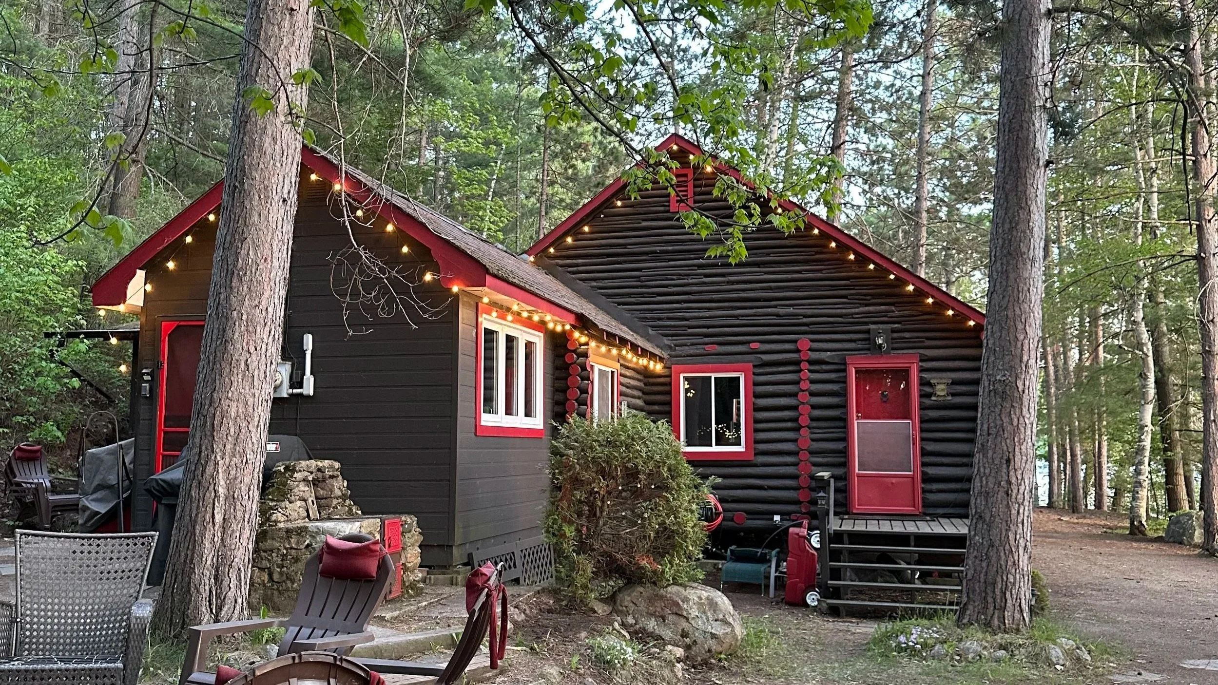 A black log cabin with red trim in a wooded area, decorated with string lights along the roofline, surrounded by trees and outdoor furniture.