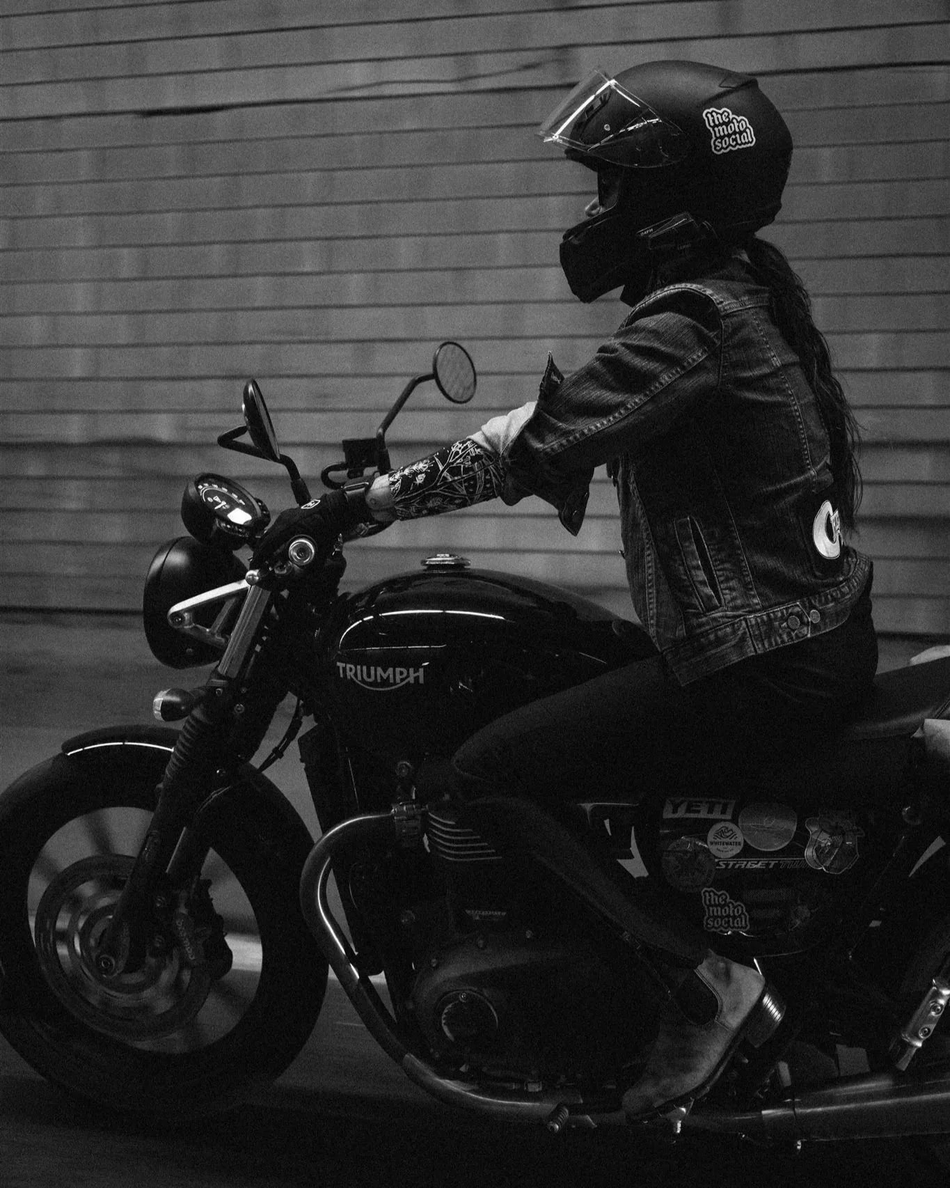A person riding a Triumph motorcycle, wearing a helmet, a denim jacket, and gloves, in front of a wooden wall, in black and white.