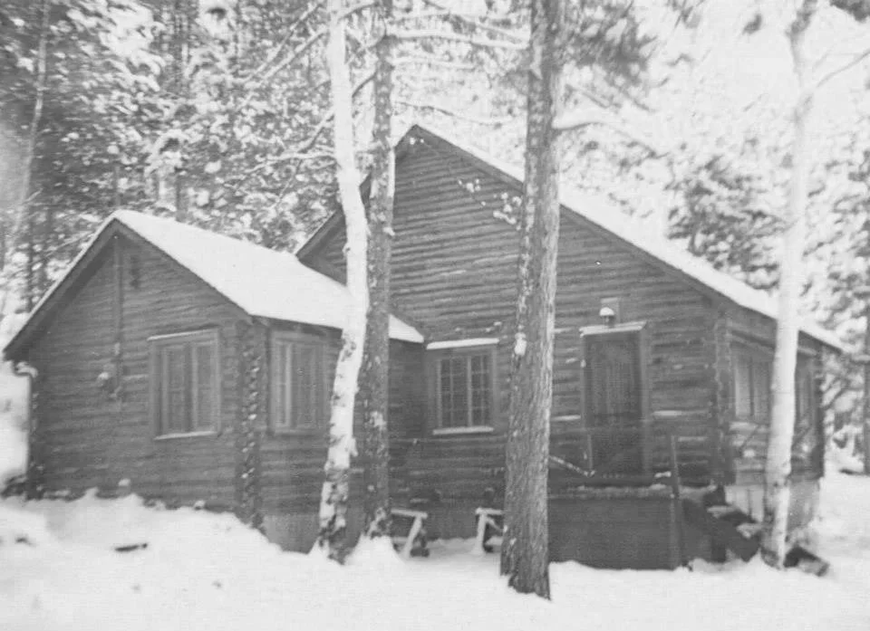 A black and white photo of a wooden cabin in a snowy forest with tall trees surrounding it.