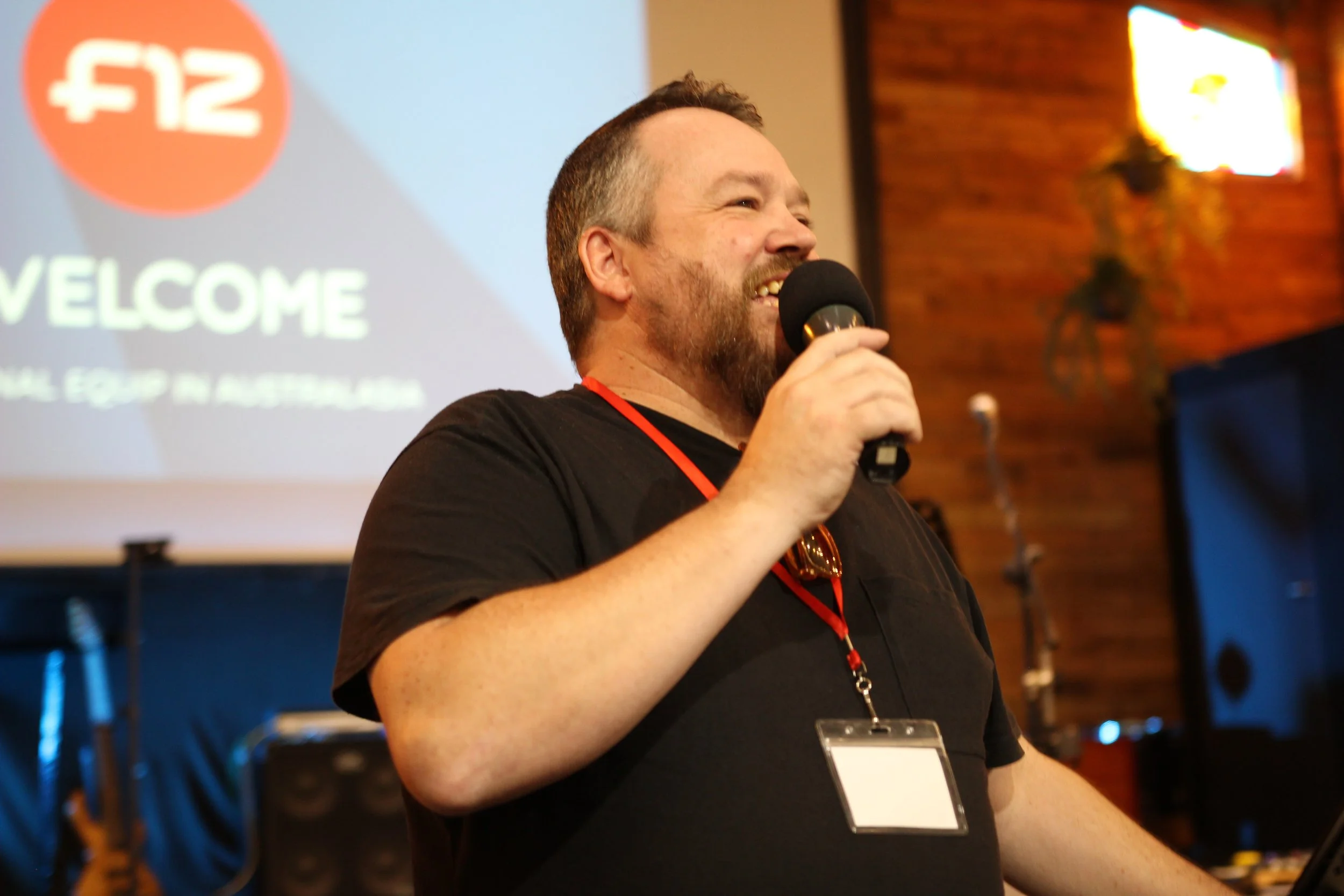 Image is of a smiling man in black tshirt with a microphone welcoming people to the 2025 Four12 Australasia Equip. There is a projection on the wall behind him with Welcome and the Four12 logo.