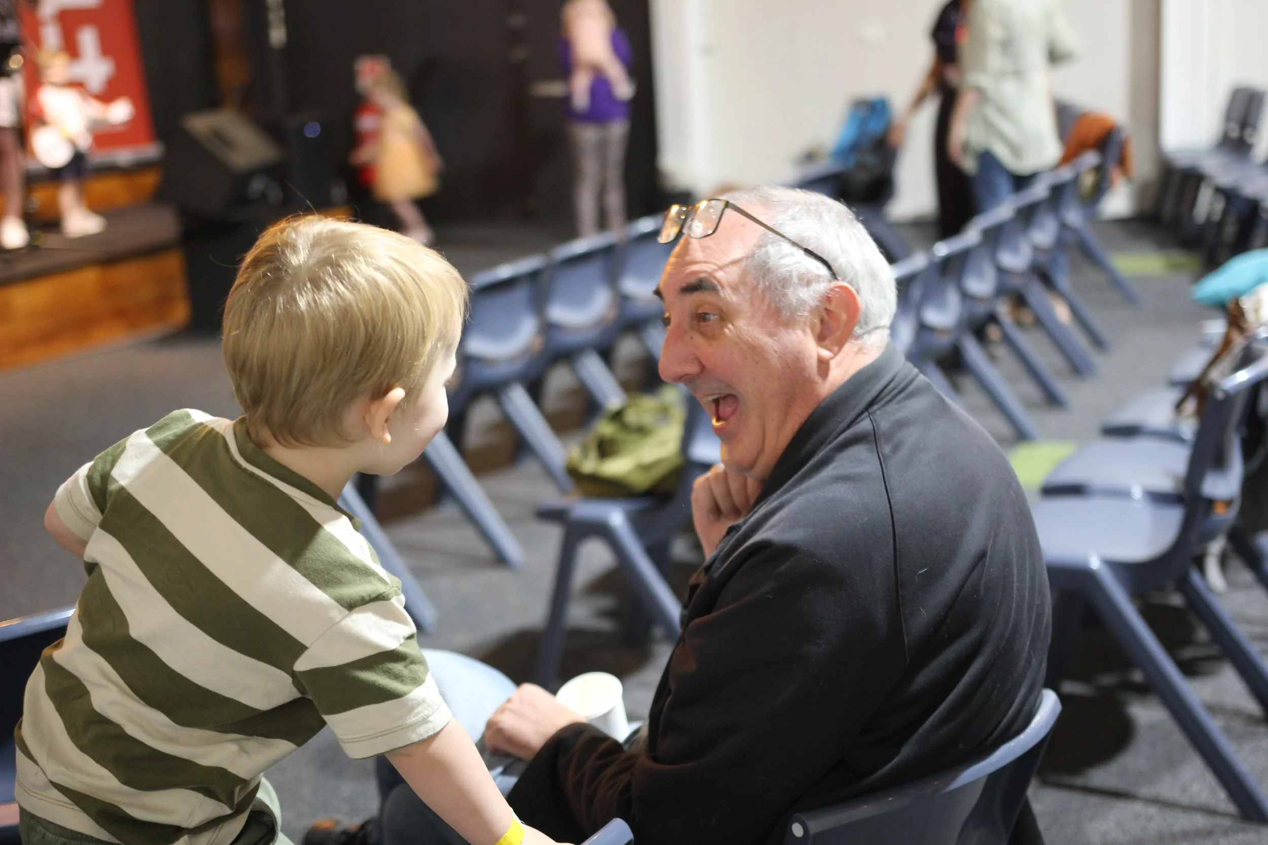 Man and small boy having fun and enjoying each other's company. Man is sitting in a row of chairs and has a big smile on his face as he interacts with boy. Boy has a striped shirt and is standing on a chair facing the man and leaning in smiling