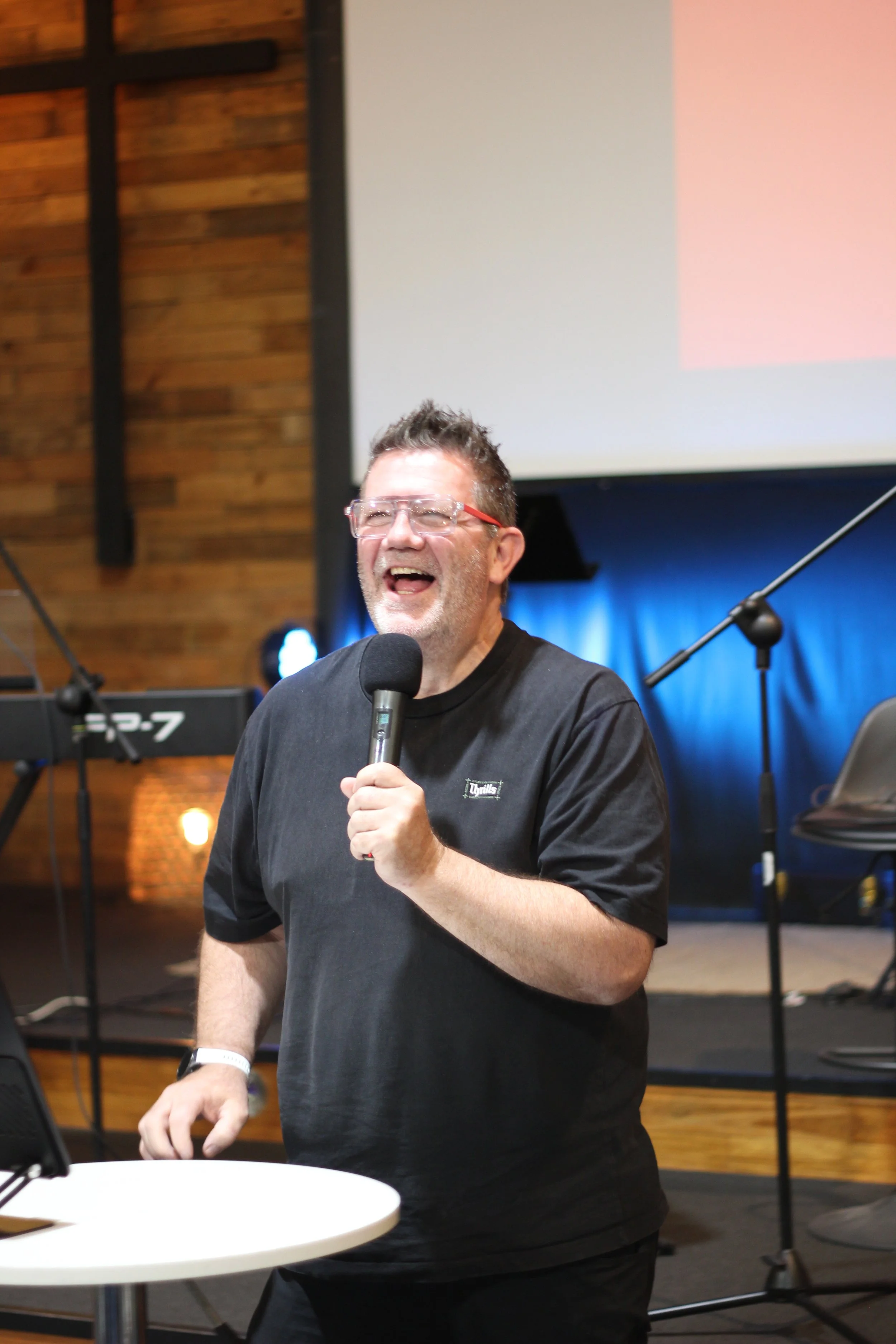 A man in a black tshirt holding a microphone in front of a stage. He has a huge smile on his face and is full of joy sharing about Jesus