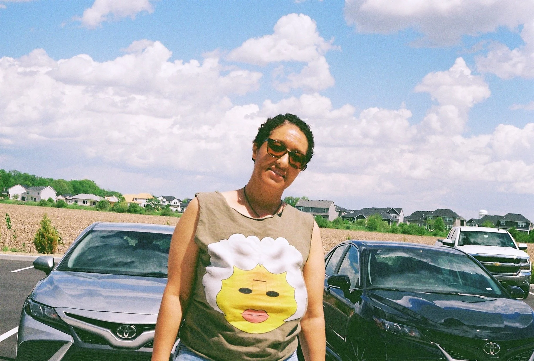 A woman with short curly hair, wearing sunglasses and a sleeveless T-shirt with a cartoon face, standing in front of parked cars in an outdoor parking lot with houses and fields in the background.