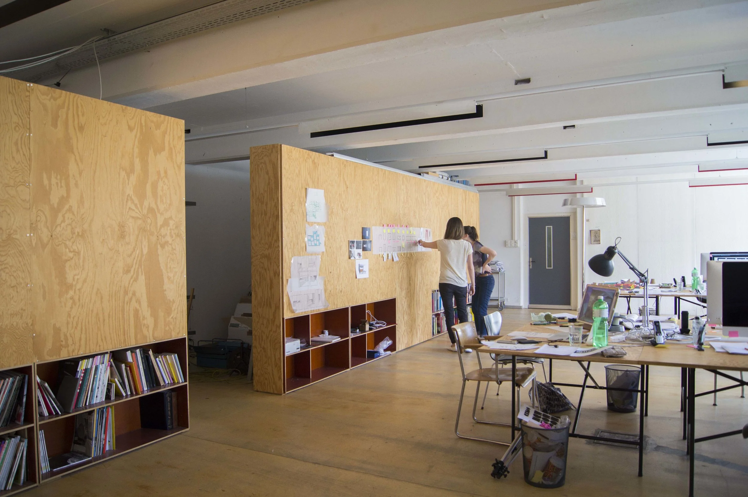 Ein modernes Büro mit Holzelementen, Arbeitstischen, Stühlen, Computern und einer weißen Wand. Zwei Frauen arbeiten an einer Wand mit Papieren und Notizen. Es gibt Bücherregale, einen Tisch mit Arbeitsmaterialien, Wasserflaschen und einen Schreibtisch mit Computer und Schreibwaren.