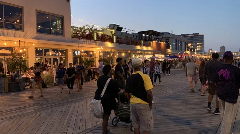 People walking along a wooden boardwalk outside a bustling waterfront restaurant and shops with string lights and outdoor seating during early evening.