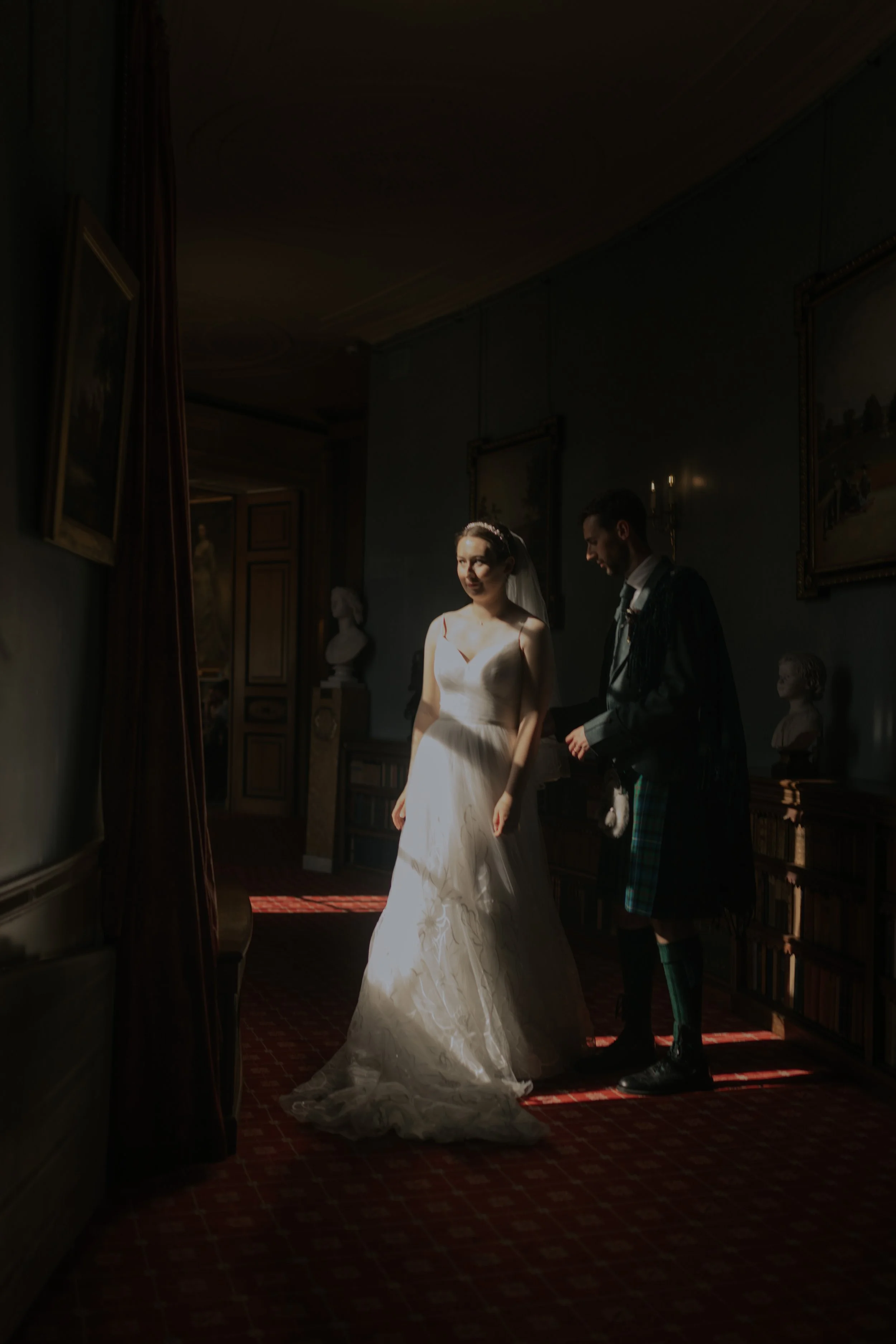 A bride in a white wedding gown and veil standing beside a groom in a kilt, inside a dimly lit room with paintings on the walls and sculptures on shelves.