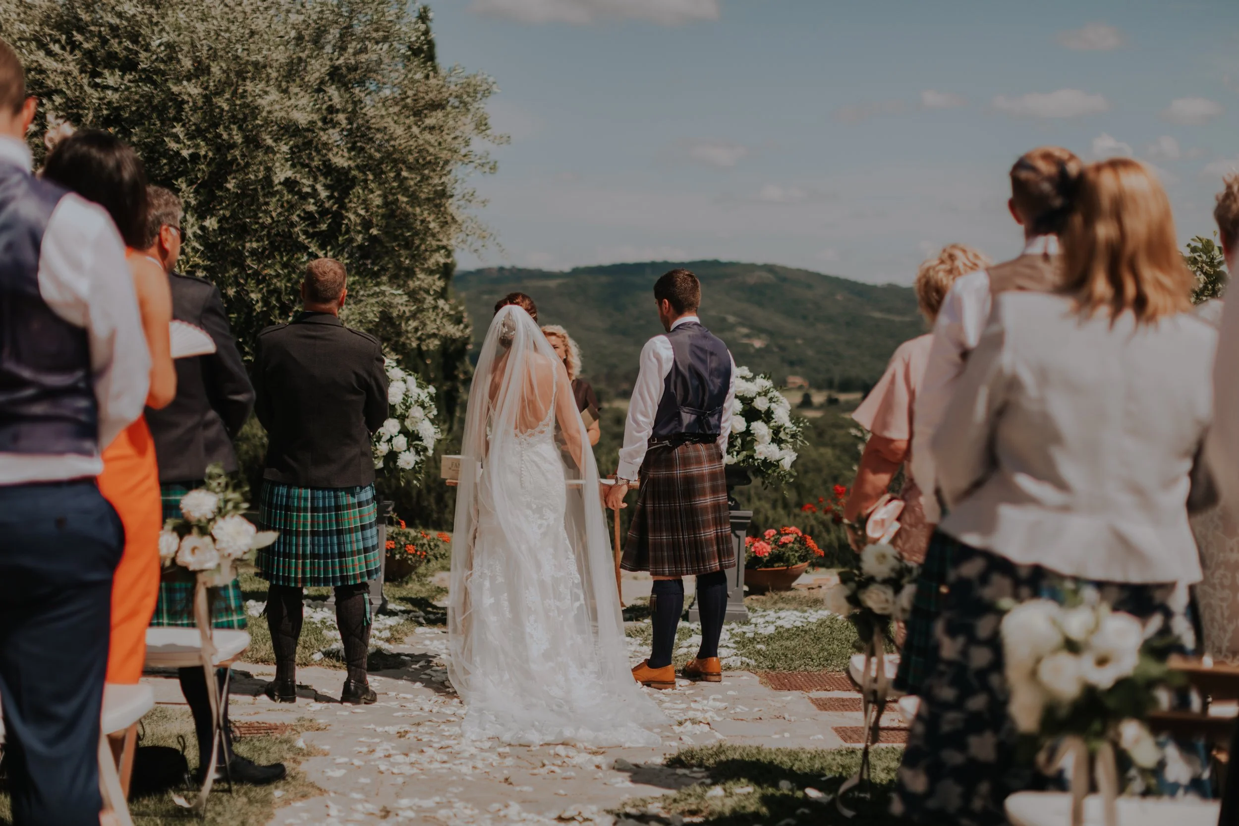 A wedding ceremony outdoors with the bride and groom holding hands in front of an officiant, surrounded by guests, green trees, and mountains in the background on a sunny day.