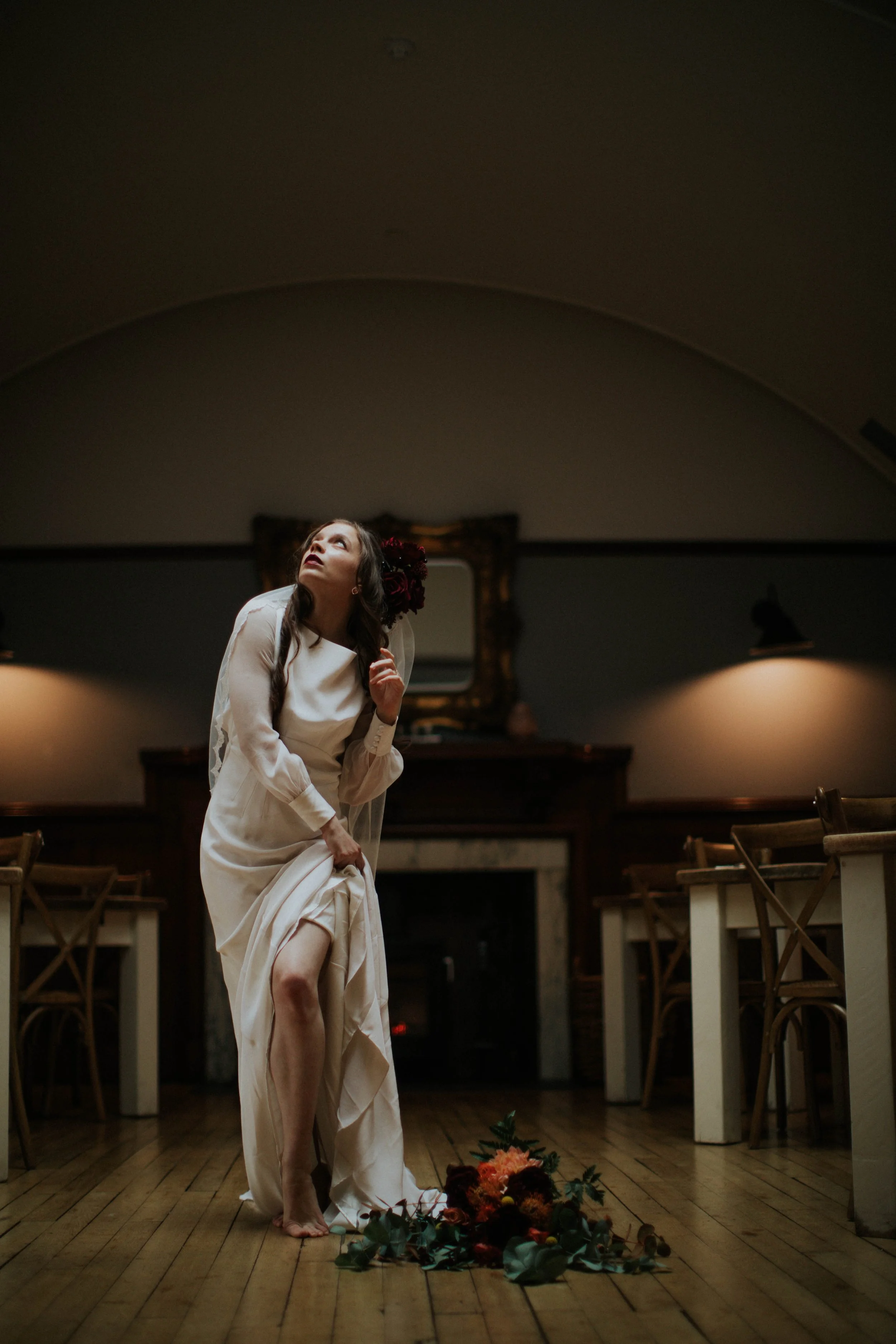 A woman in a white dress stands barefoot on a wooden floor, looking upward, with a bouquet of flowers on the floor in front of her. The room has dim lighting, a mirror on the back wall, and wooden chairs around.