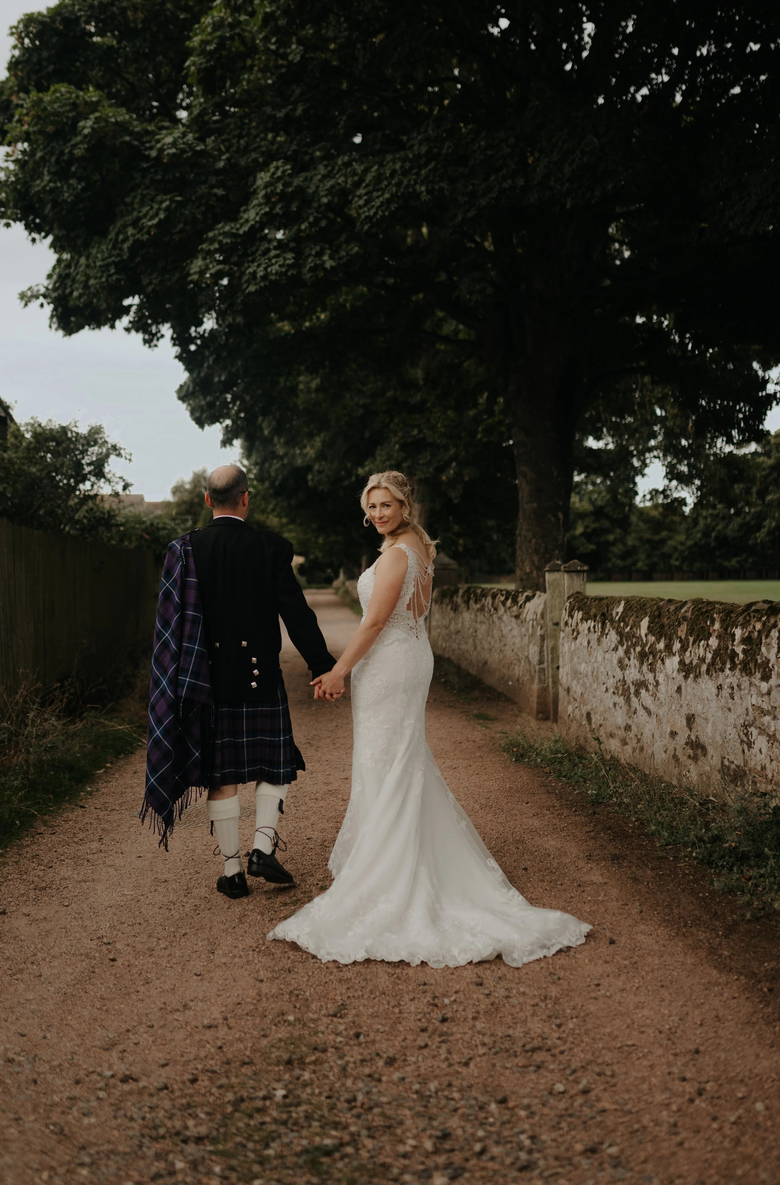 Lori and Chris walking hand in hand along a tree-lined path beside a stone wall, with Lori looking back in her lace wedding dress.
