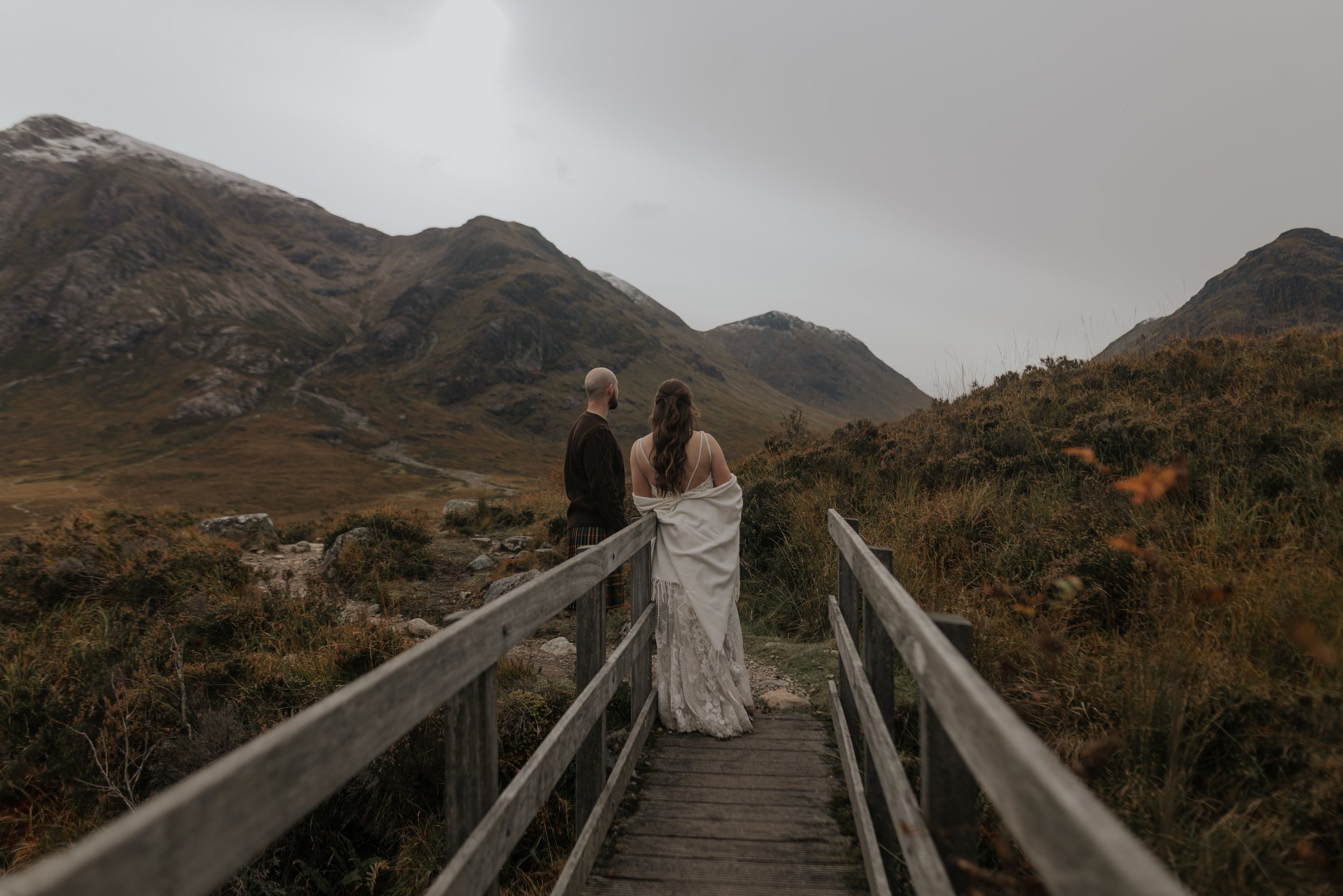 A couple standing on a wooden footbridge overlooking a scenic mountain landscape with cloudy skies.