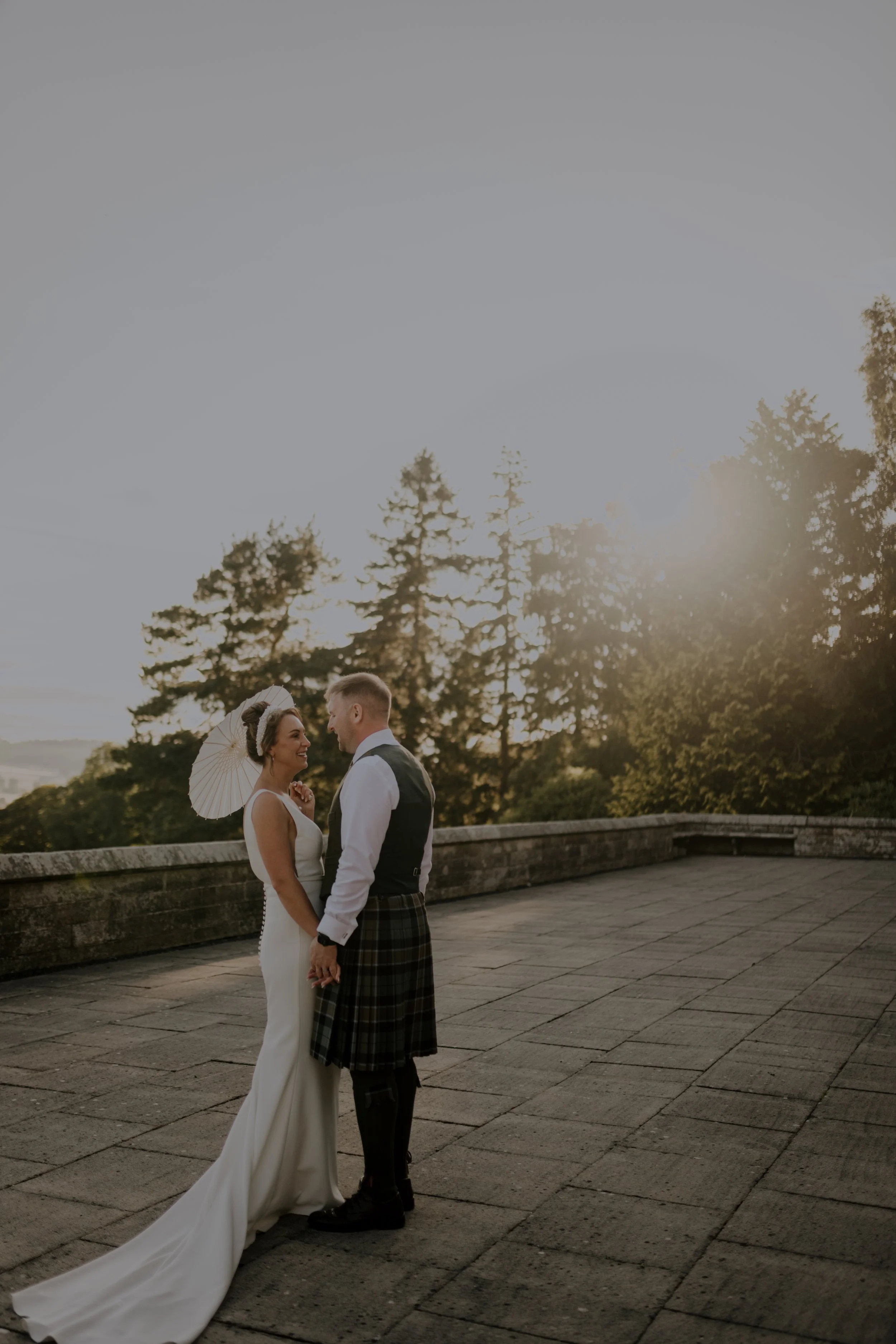 A bride and groom holding hands and facing each other on a stone terrace during sunset, with trees and the sky in the background.