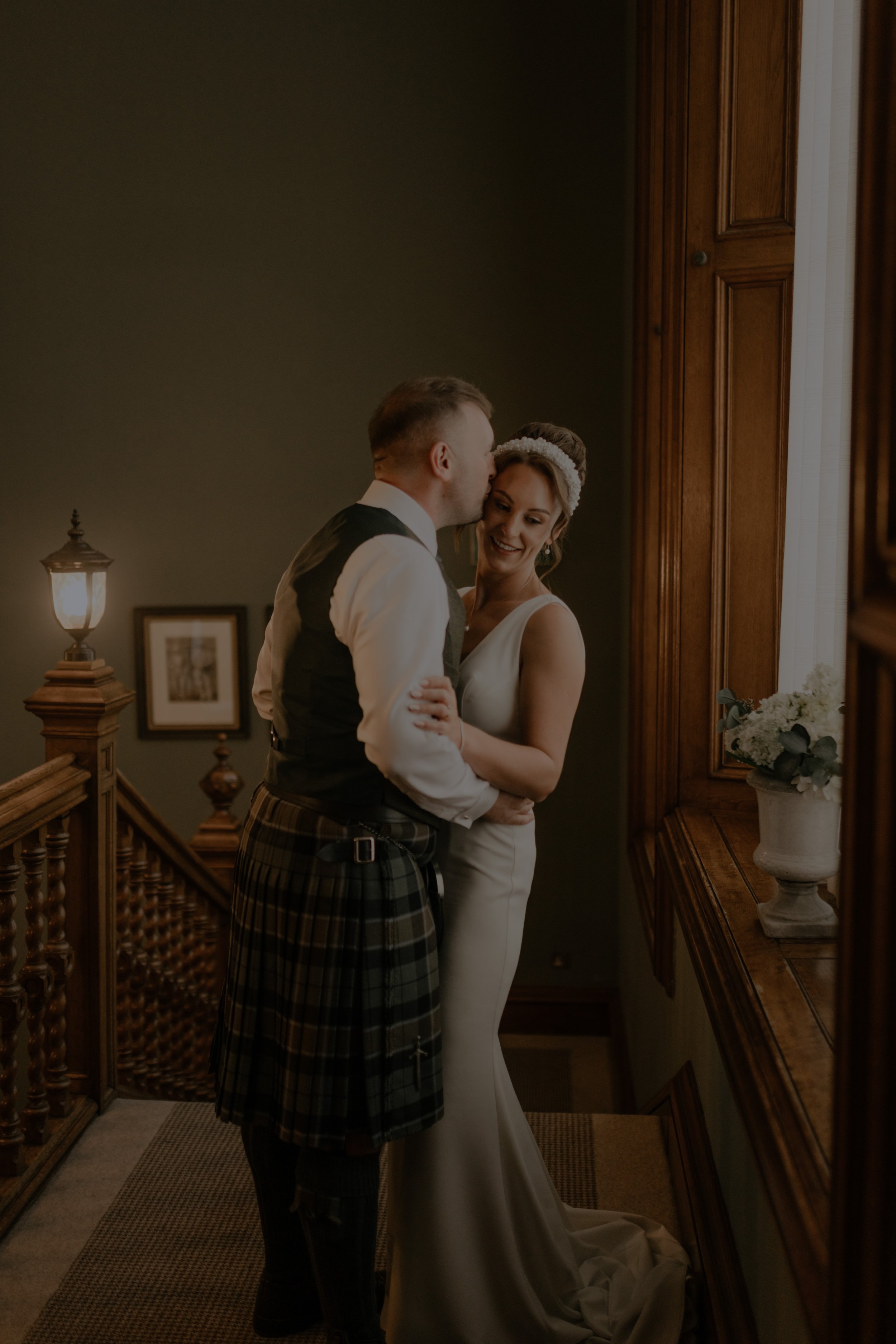 A bride and groom sharing a tender moment indoors, with the groom kissing the bride on the forehead near a wooden window.