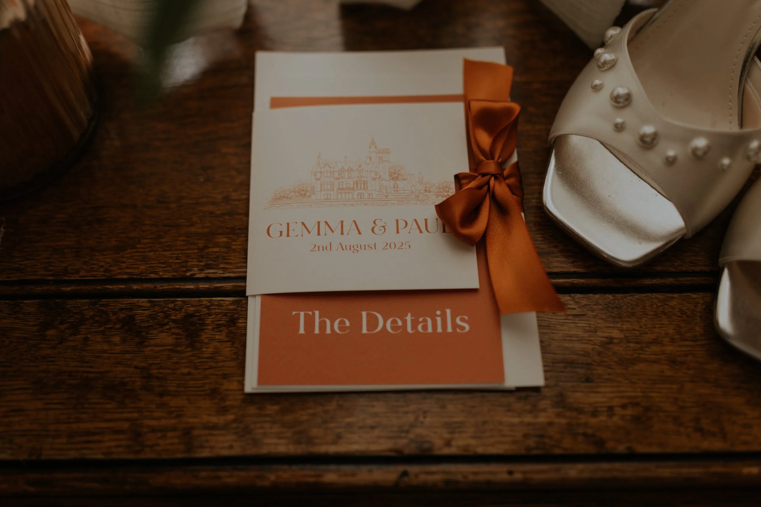 Wedding invitation card with the names Gemma & Paul, date August 2, 2025, and a ribbon bow, placed on a wooden surface near a pair of white high-heeled shoes.