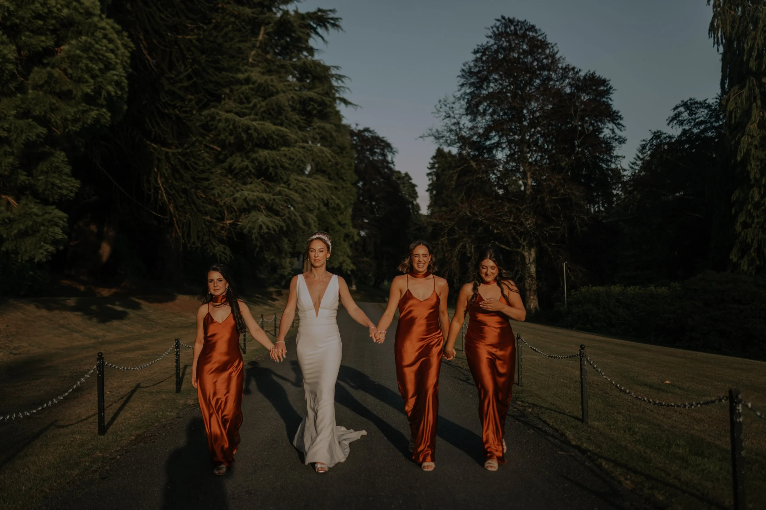 Four women in formal dresses holding hands and walking on a path surrounded by trees during sunset.