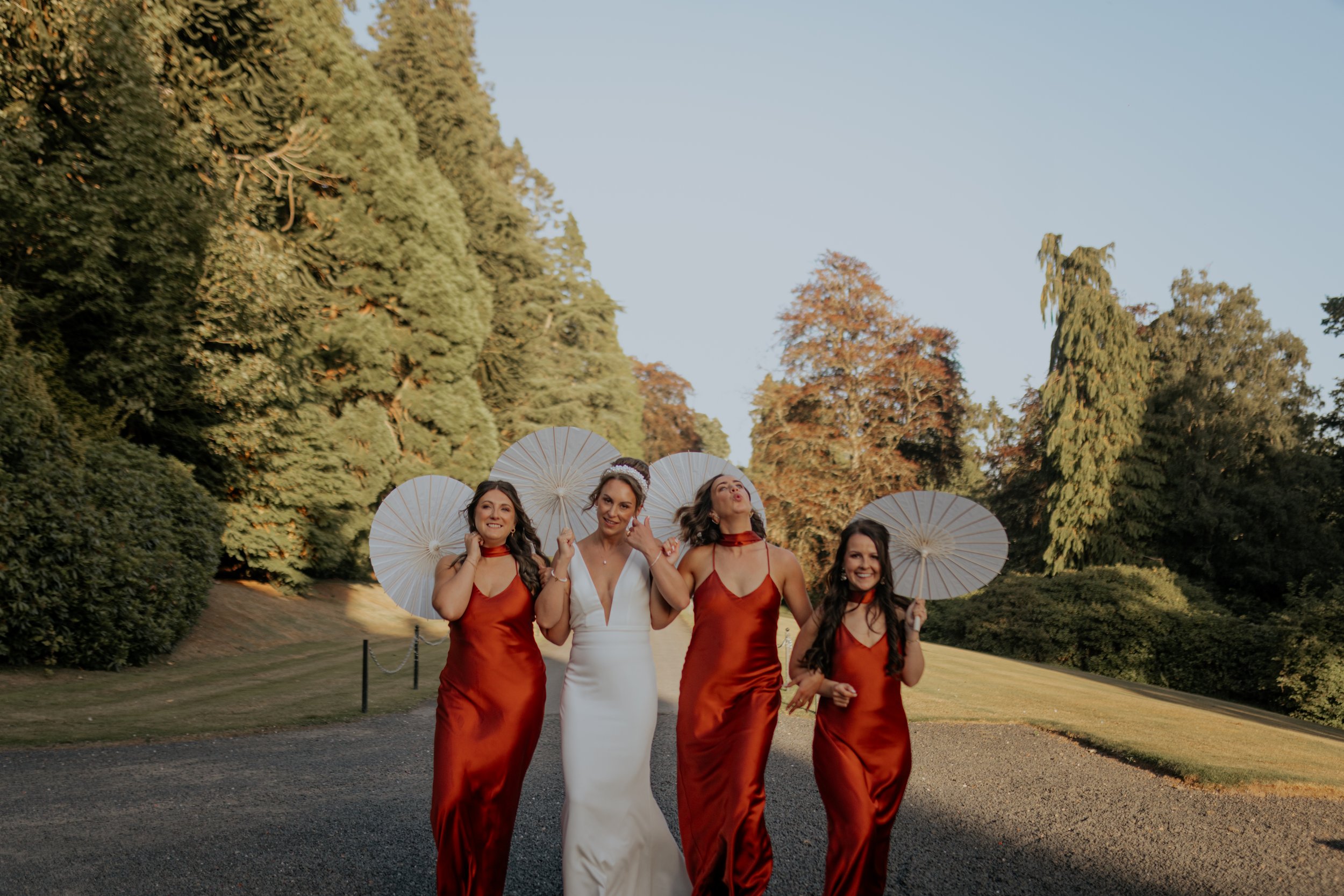 A bride and three bridesmaids walking outdoors on a gravel path, each holding a white parasol, with green trees in the background. The bride is wearing a white dress and the bridesmaids are in matching red satin dresses.