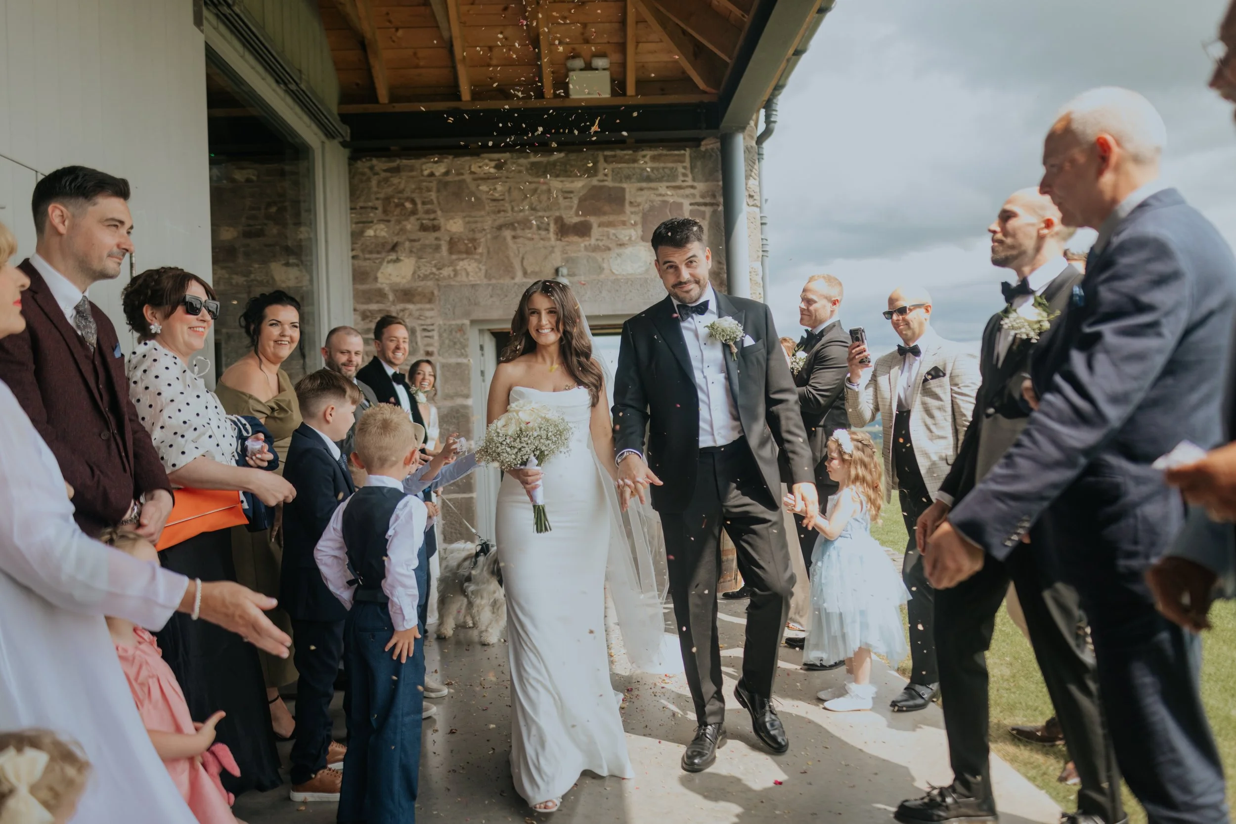 Bride and groom walking hand in hand through a crowd of wedding guests outside a stone building, with guests throwing confetti, during a wedding celebration on a partly cloudy day.