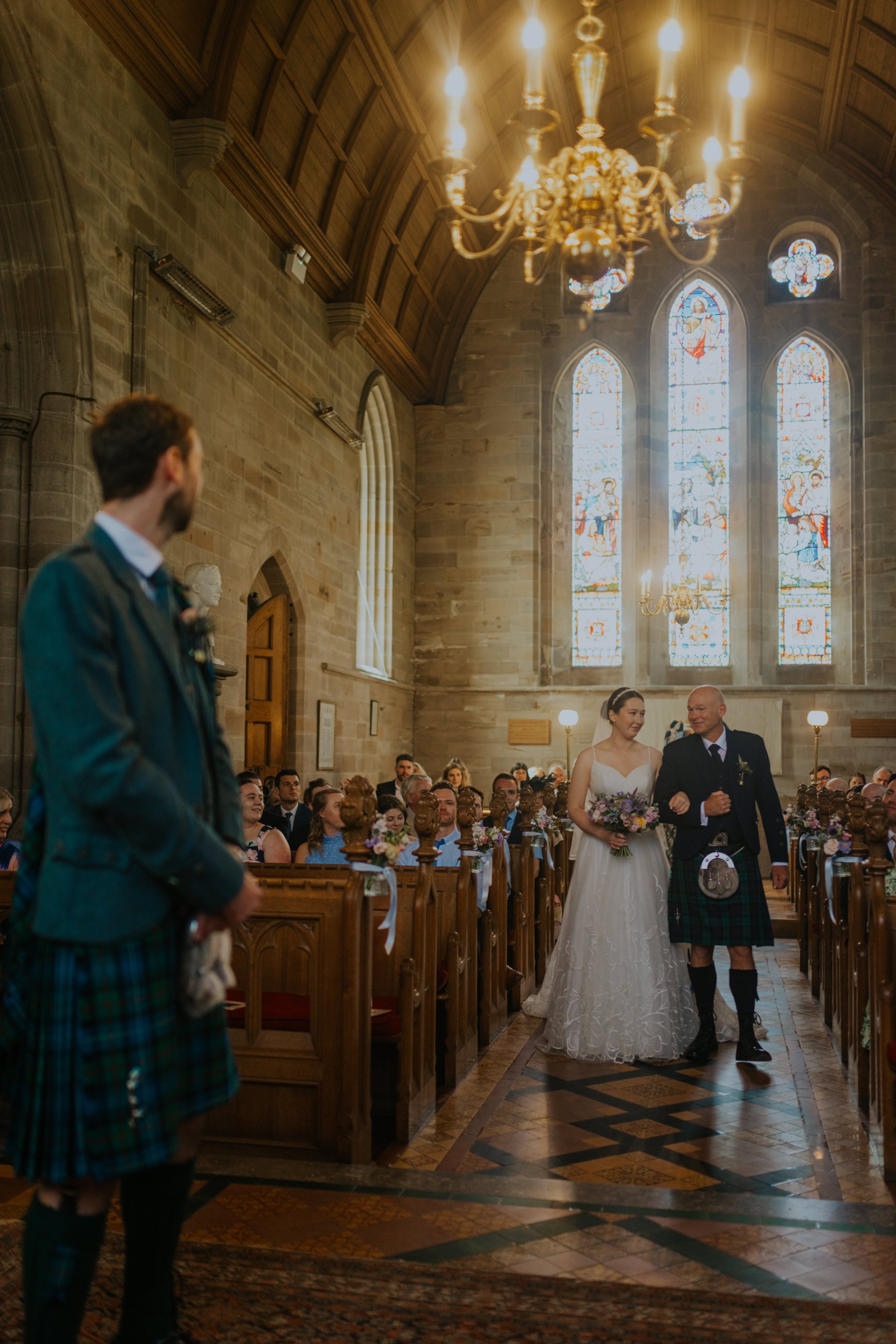 A bride walking down the aisle with a man in a kilt during a wedding ceremony inside a church with stained glass windows.
