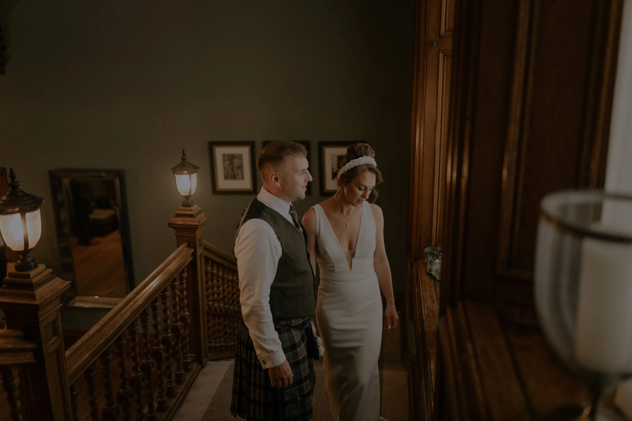 A man in formal attire and a woman in a white dress standing on a staircase landing, looking down. The staircase features wooden railing, and the background includes framed pictures and warm lighting.