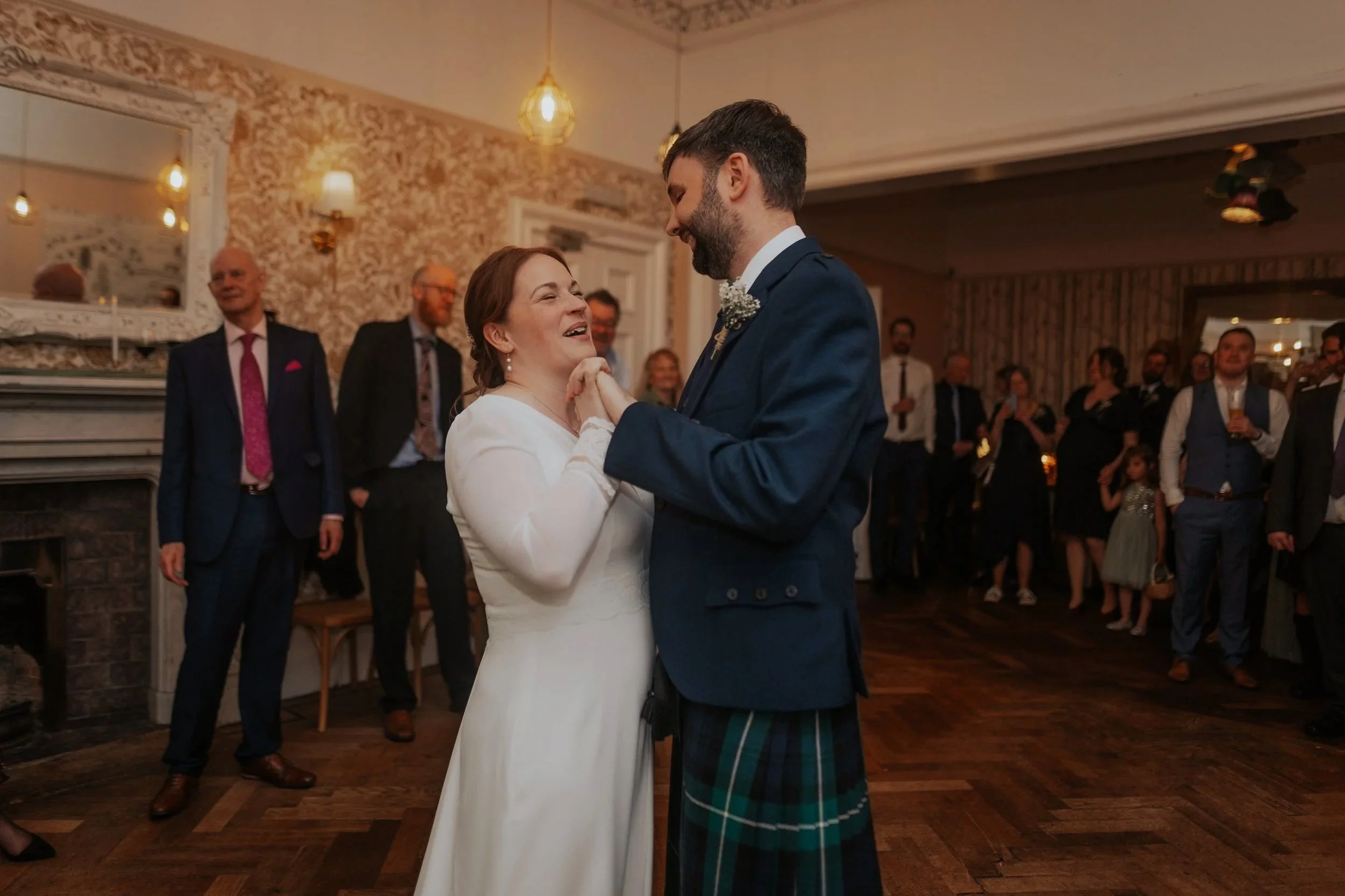 A bride and groom dance together at their wedding reception, surrounded by guests in a warmly lit, decorated room.