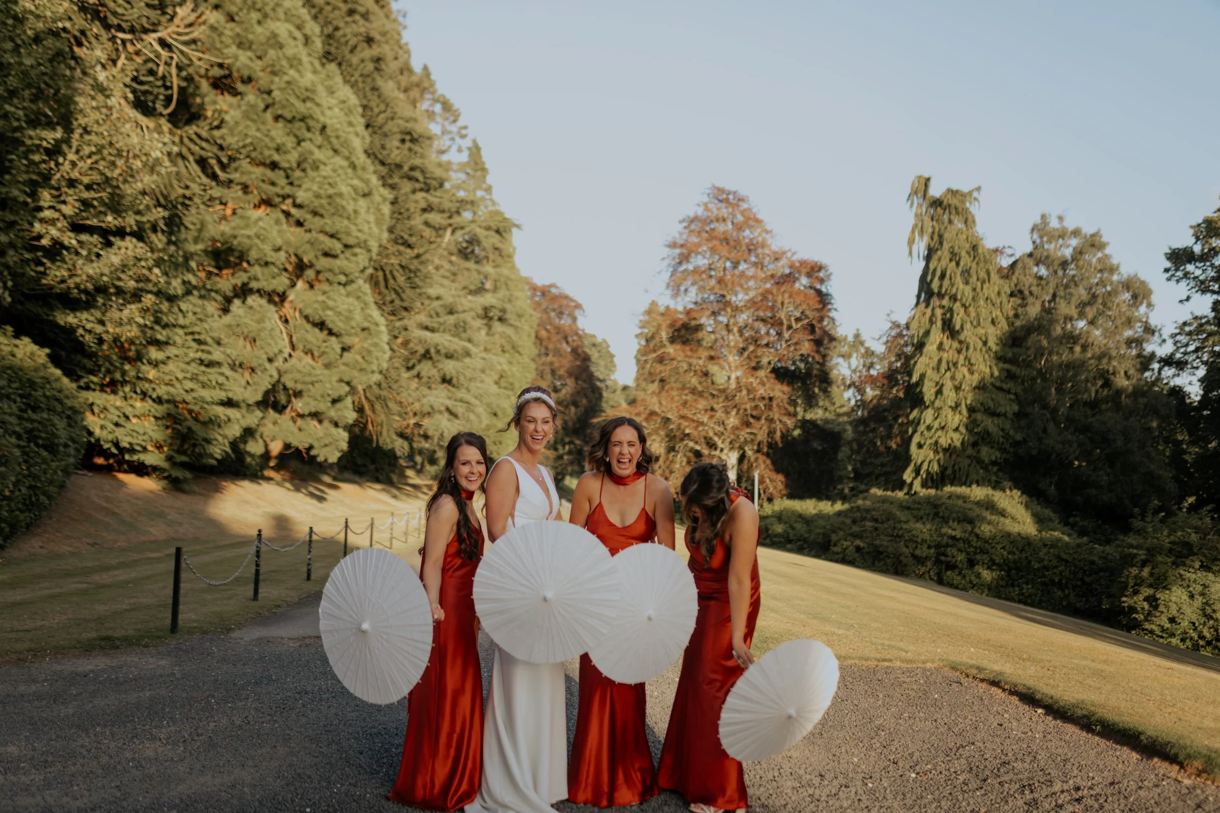 Four women dressed in formal gowns, two in white and two in red, standing outdoors with umbrellas, smiling and laughing, surrounded by large trees and greenery.
