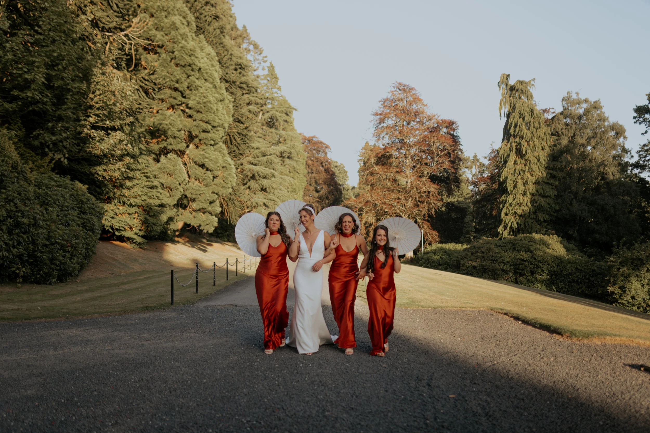 A bride in a white wedding dress walking with four bridesmaids in red dresses, holding white parasols, walking on a gravel path in a park with trees and greenery during daytime.