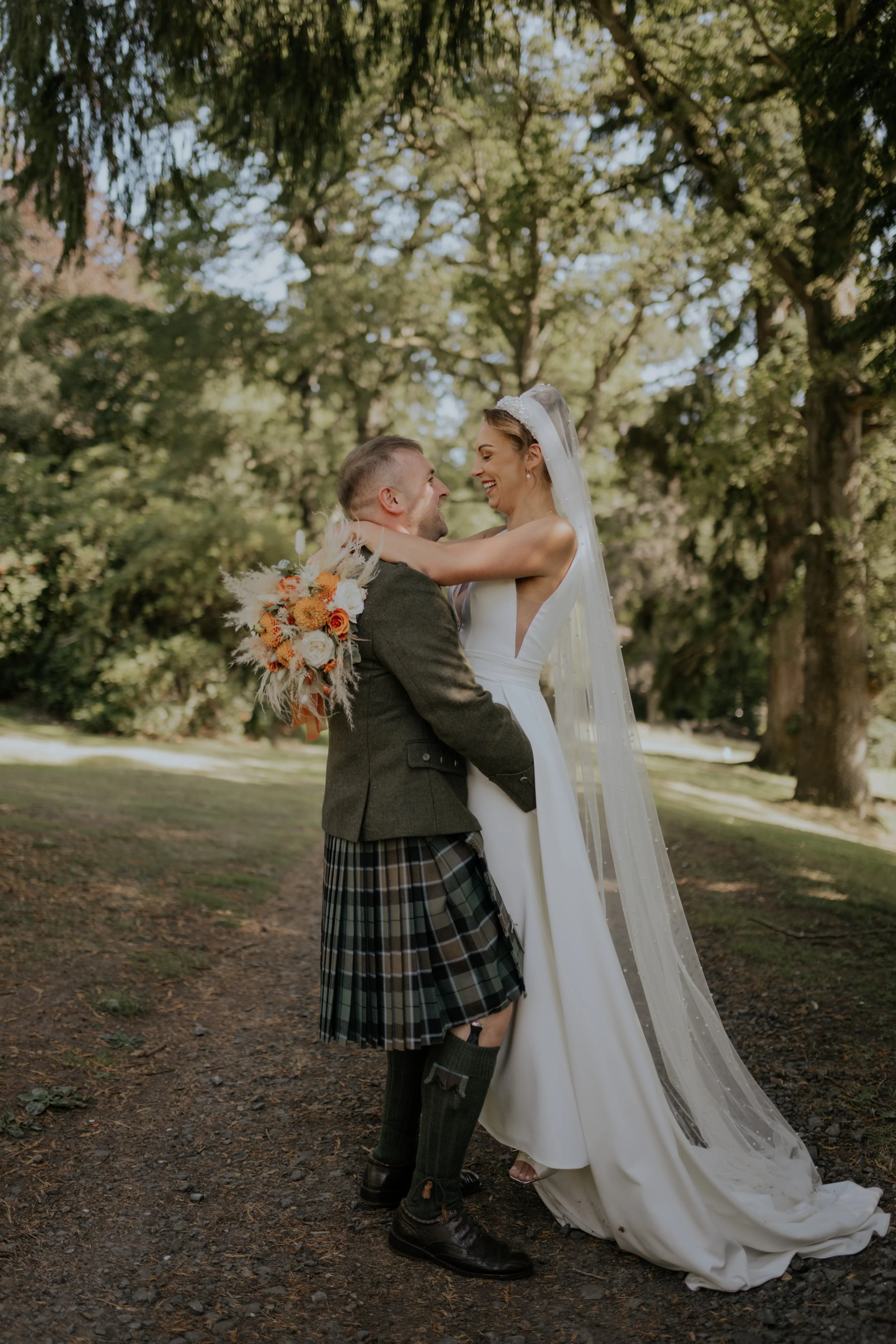 A bride and groom are smiling and embracing outdoors in a wooded area with lush green trees and sunlight filtering through.