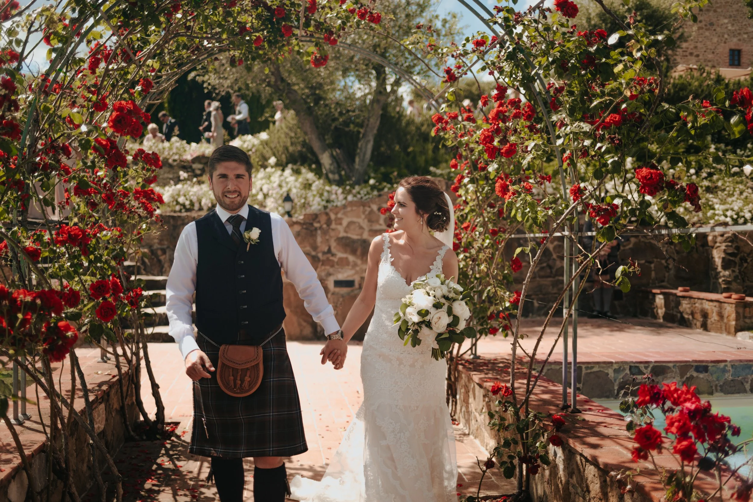 A bride and groom holding hands, walking under a floral archway on their wedding day. The bride is wearing a white lace wedding dress and holding a bouquet of white roses. The groom is dressed in traditional Scottish attire, including a kilt, waistco