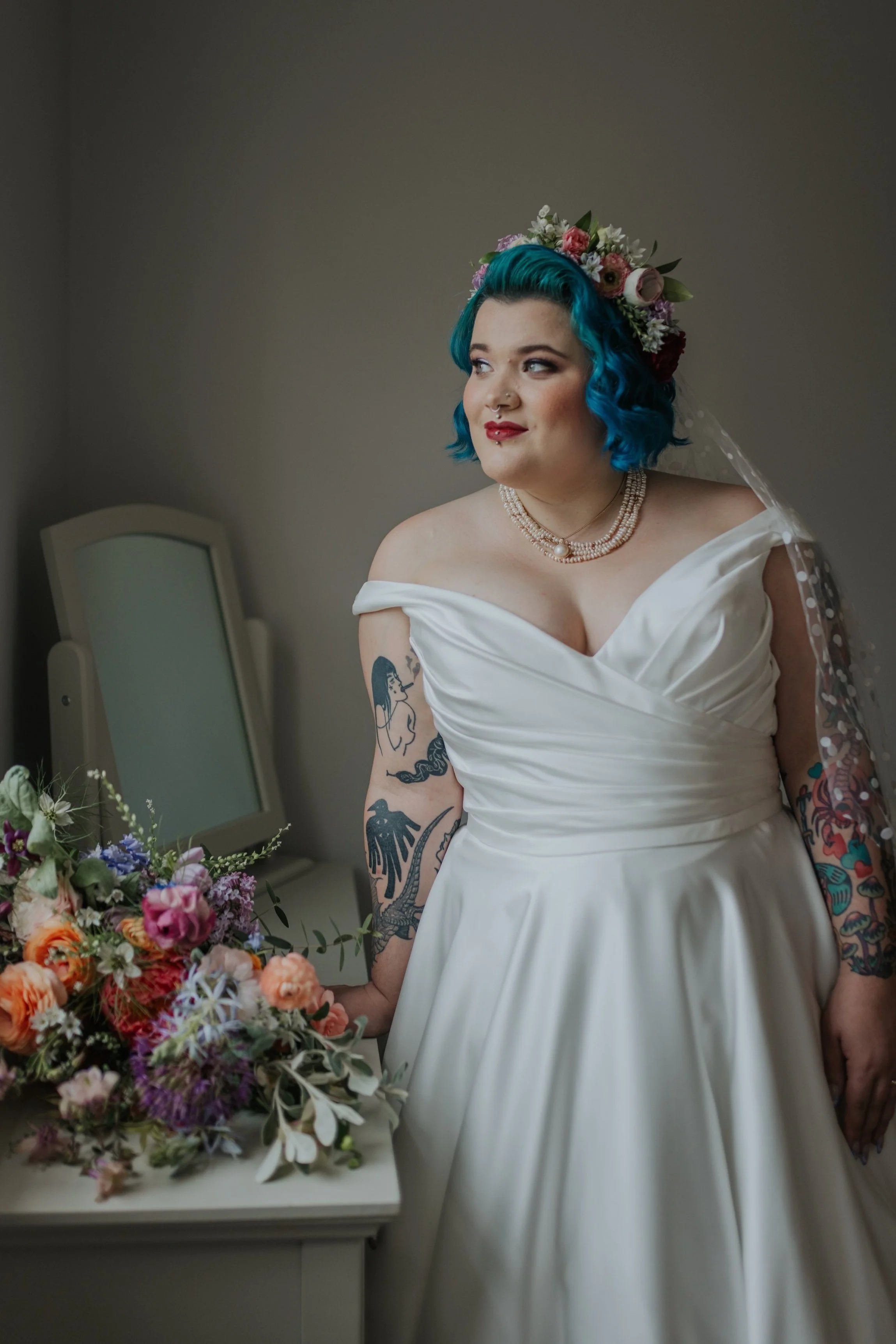 A woman with bright blue hair wearing a white off-shoulder wedding dress, a floral crown, and a pearl necklace. She has visible tattoos on her arms and stands next to a bouquet of flowers on a table.