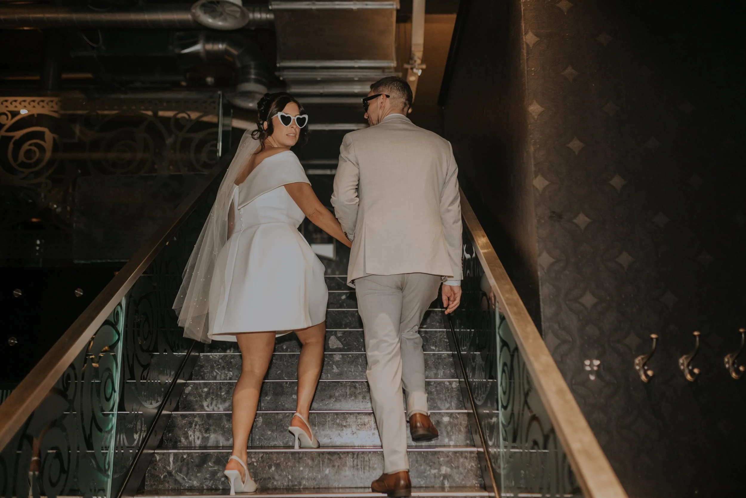 A woman in a white wedding dress and sunglasses holding hands with a man in a light-colored suit ascending a staircase.