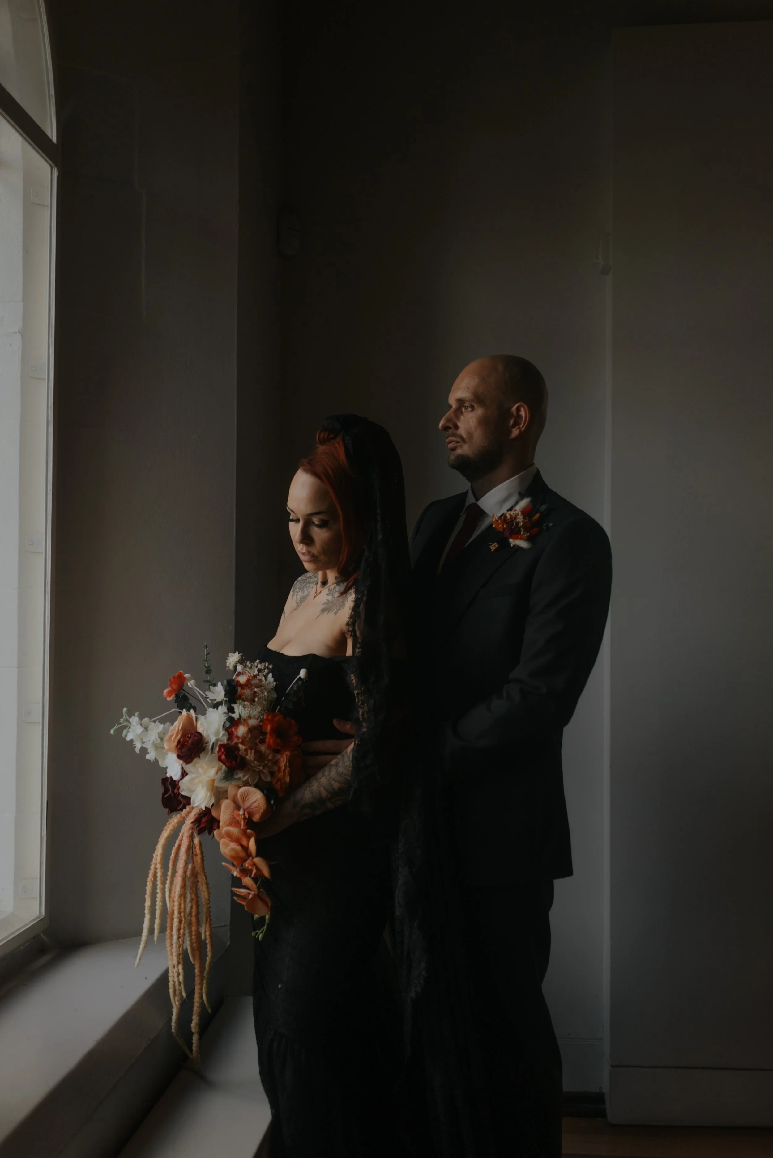 A couple dressed in formal attire standing by a window, with the woman holding a bouquet of flowers, in a dimly lit room.