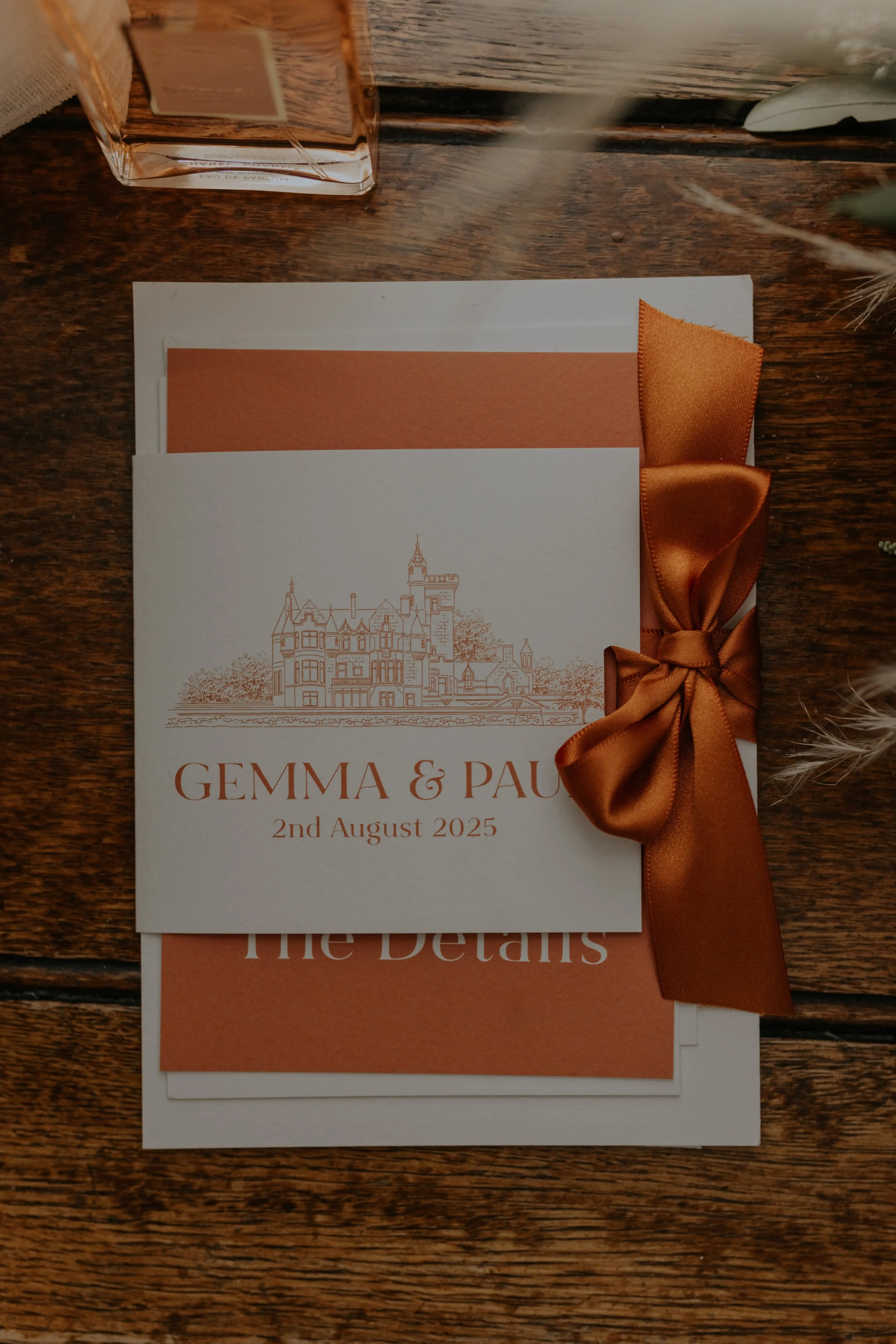 Wedding invitation card with a drawing of a castle, text with names Gemma and Paul, and the date 2nd August 2025, tied with an orange satin ribbon on a wooden surface.