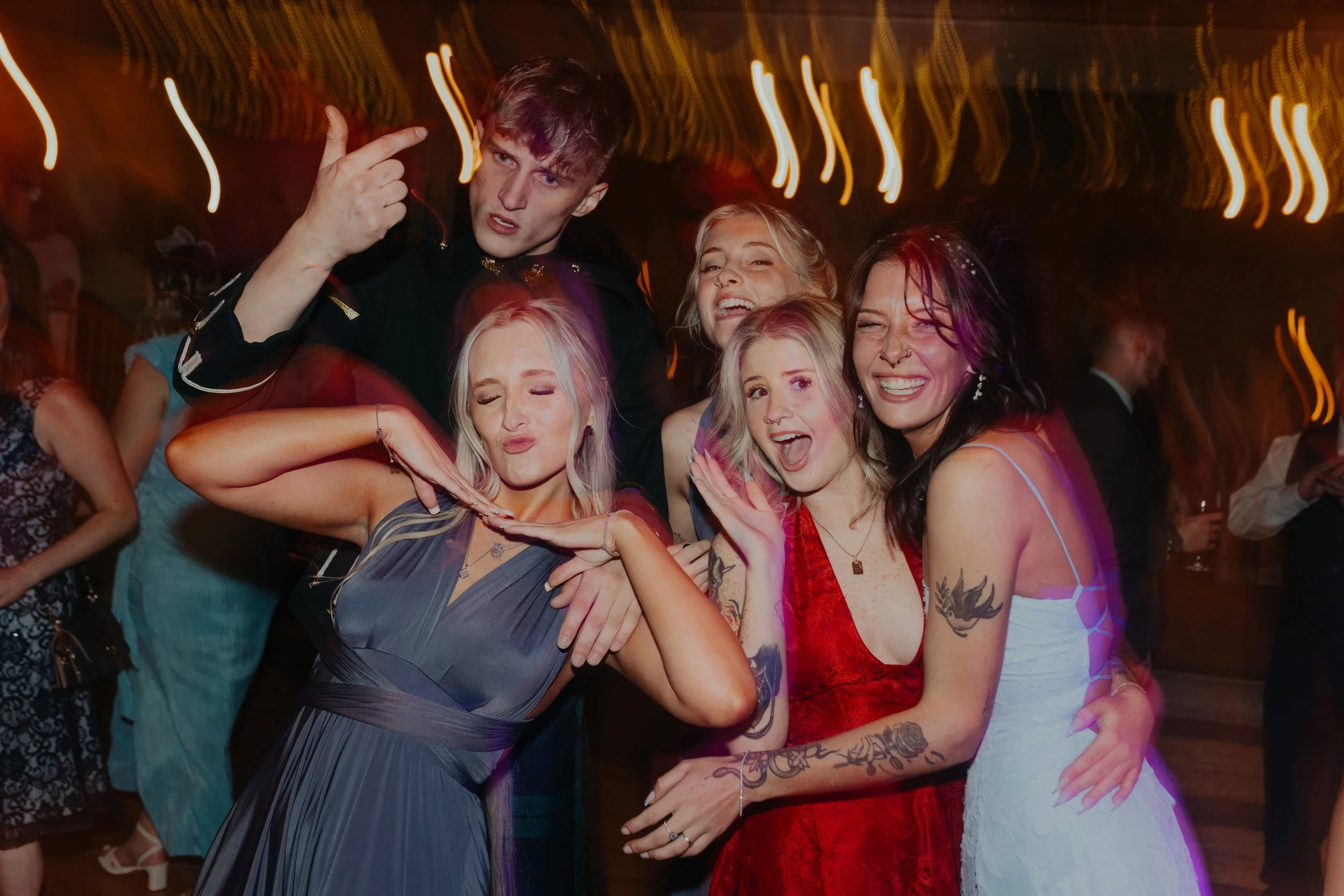 A group of six young adults celebrating at a party or wedding reception, dancing and having fun, in a dimly lit venue with colorful lights and other guests in the background.