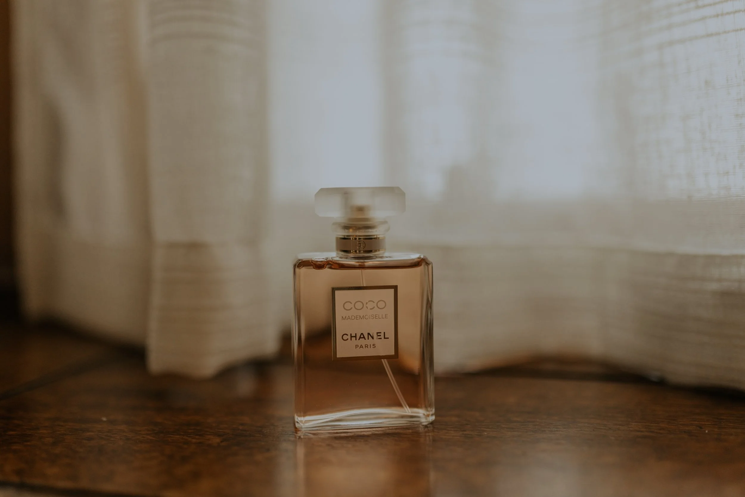 A Chanel Coco Mademoiselle perfume bottle with a pinkish hue, placed on a wooden surface near a light-colored curtain.