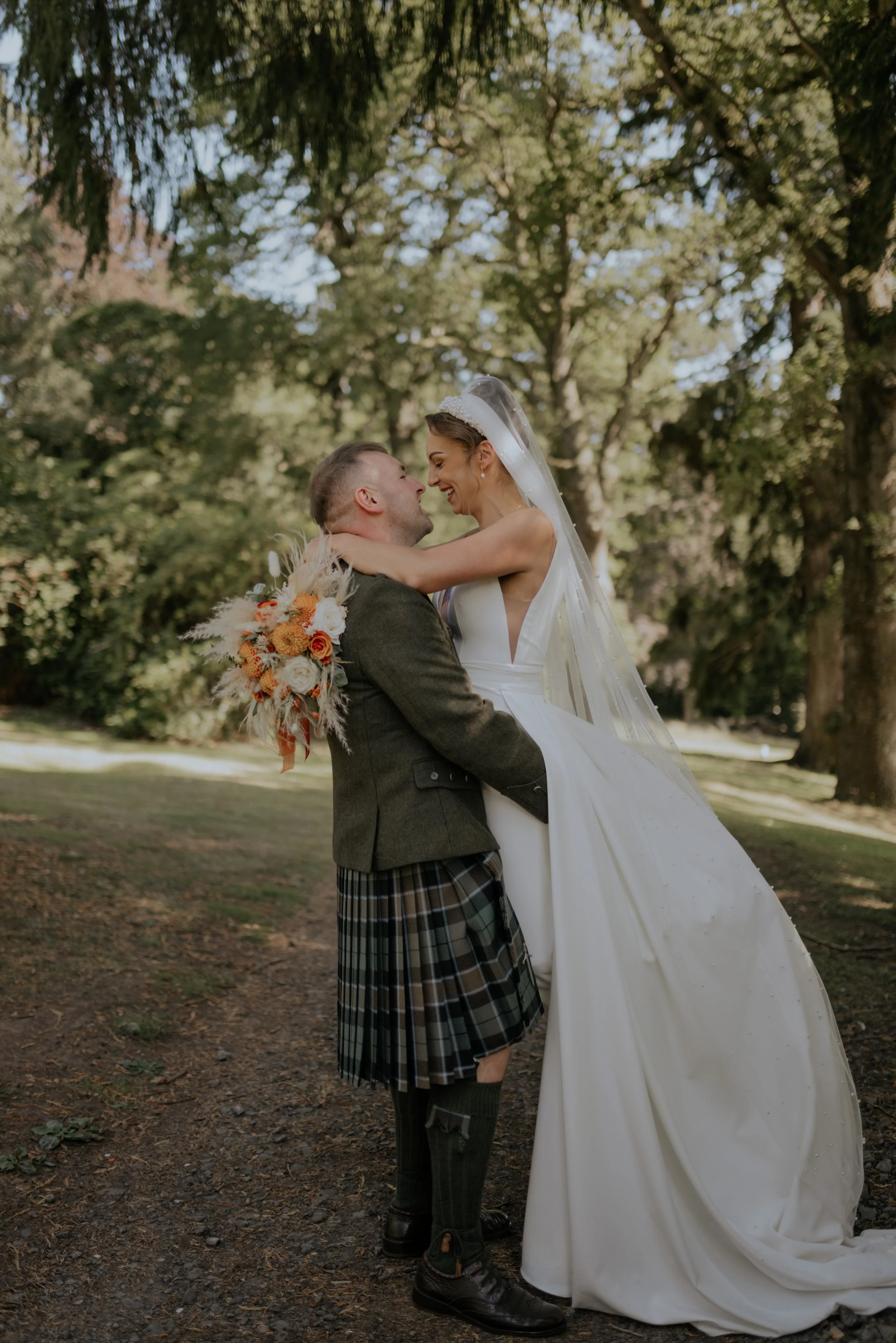 A bride and groom in wedding attire, smiling and hugging outdoors among trees.