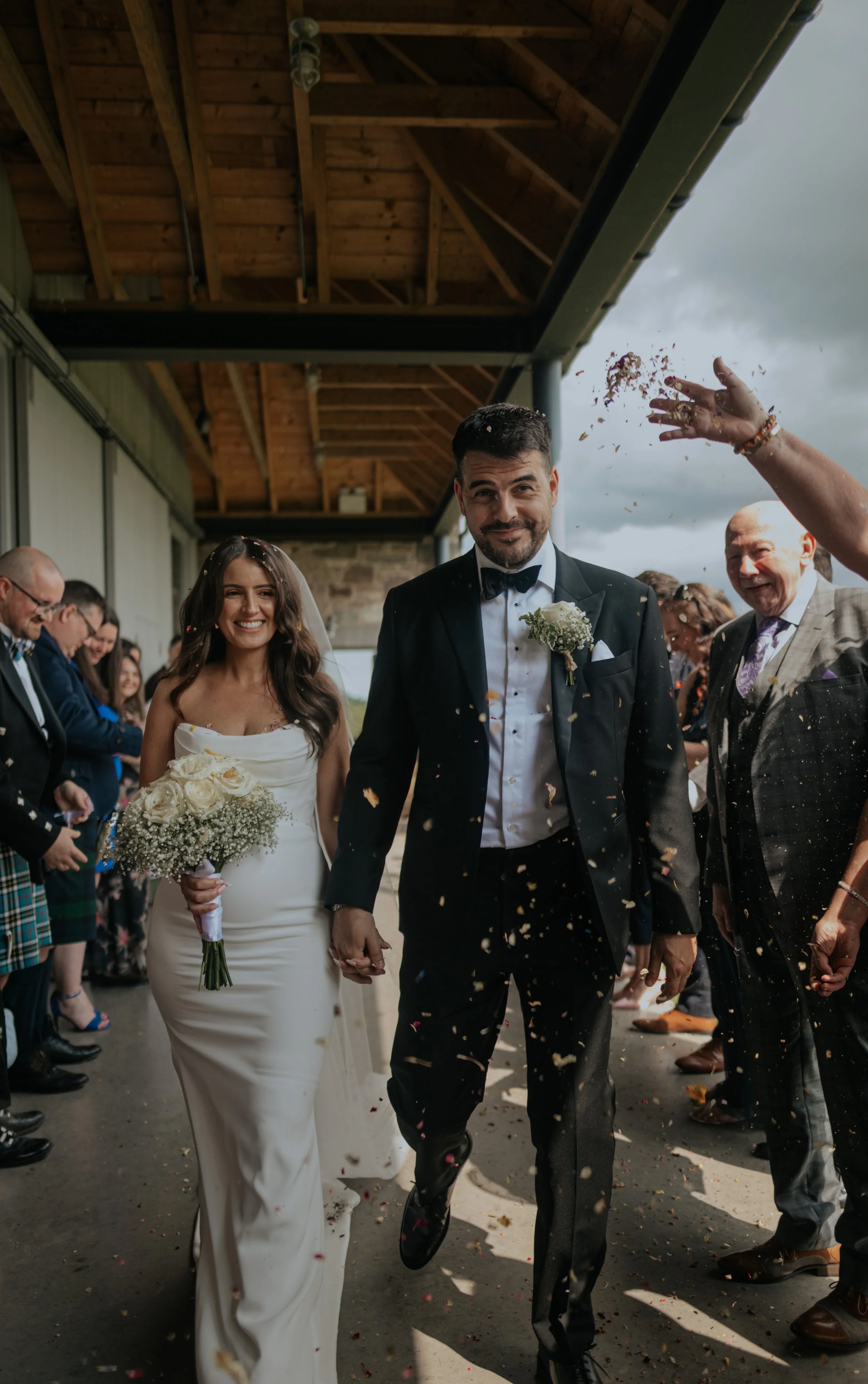 Bride and groom smiling and holding hands while walking through a crowd of wedding guests throwing confetti outside a venue with a wooden roof overhead.