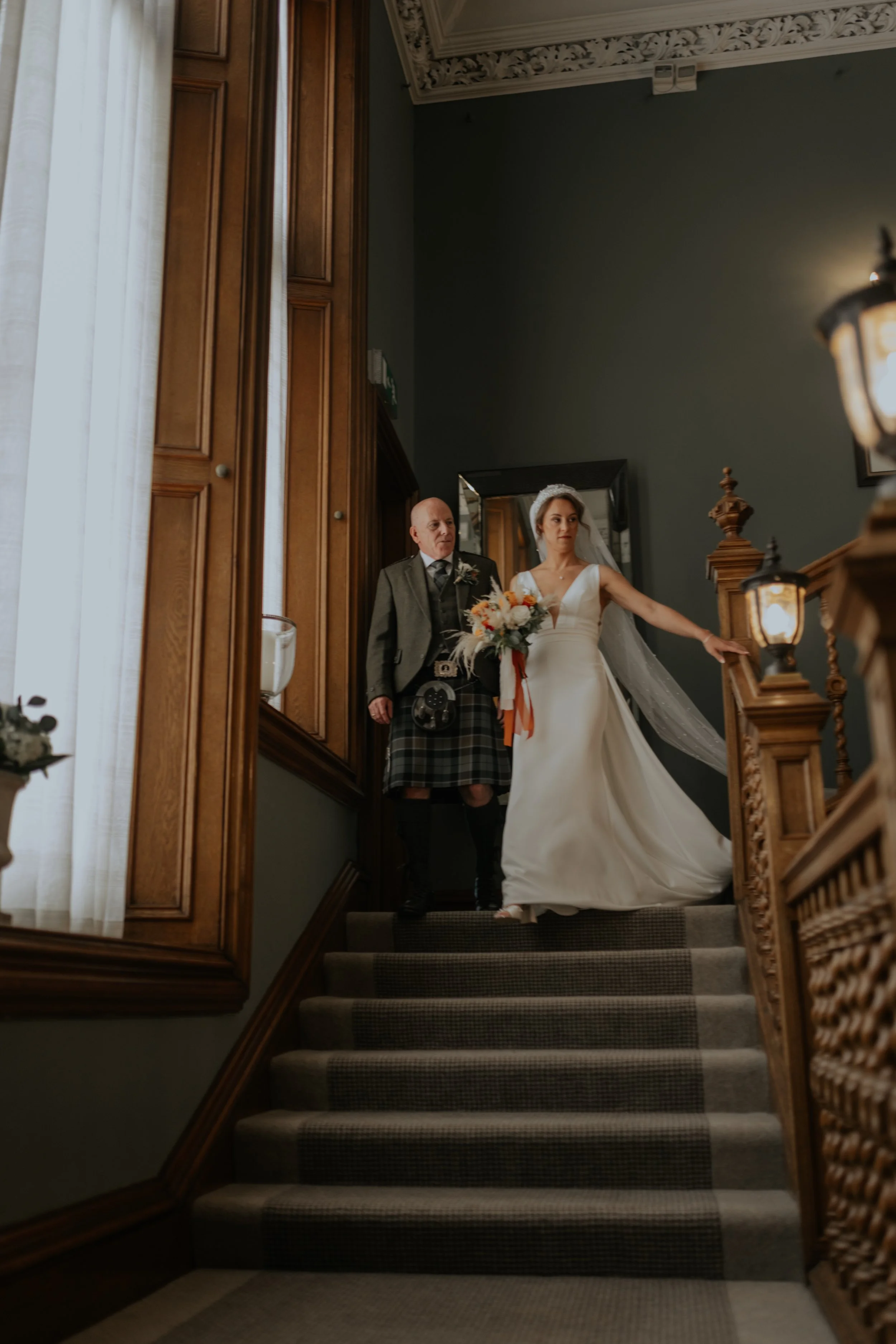 A bride in a white wedding gown and veil holding a bouquet of flowers walking down a staircase with a man in traditional Scottish attire behind her.