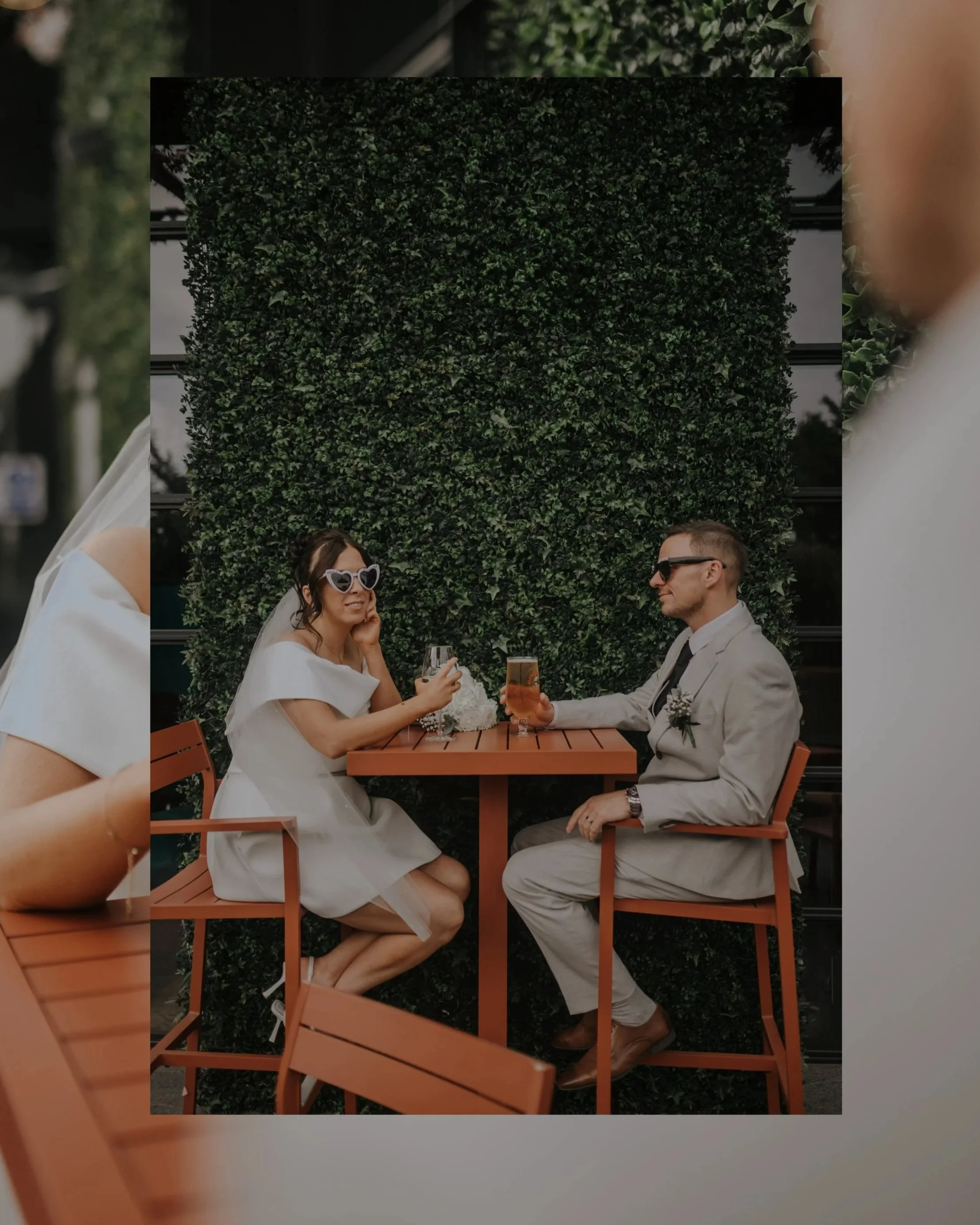 Black and white photo of a bride and groom standing in an open field, holding hands and looking down, with the bride wearing a lace wedding gown and veil, and the groom in a dark suit with a boutonniere, during sunset or dusk.