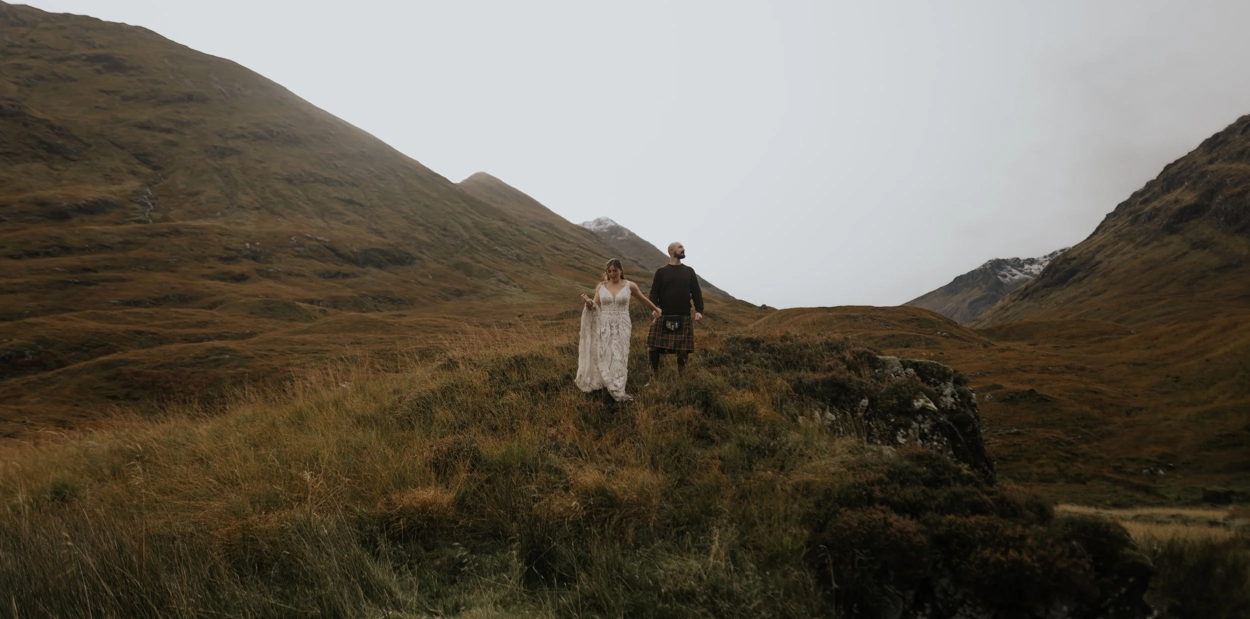 A couple in wedding attire walking hand in hand through a grassy mountain landscape, with mountains in the background under a cloudy sky.