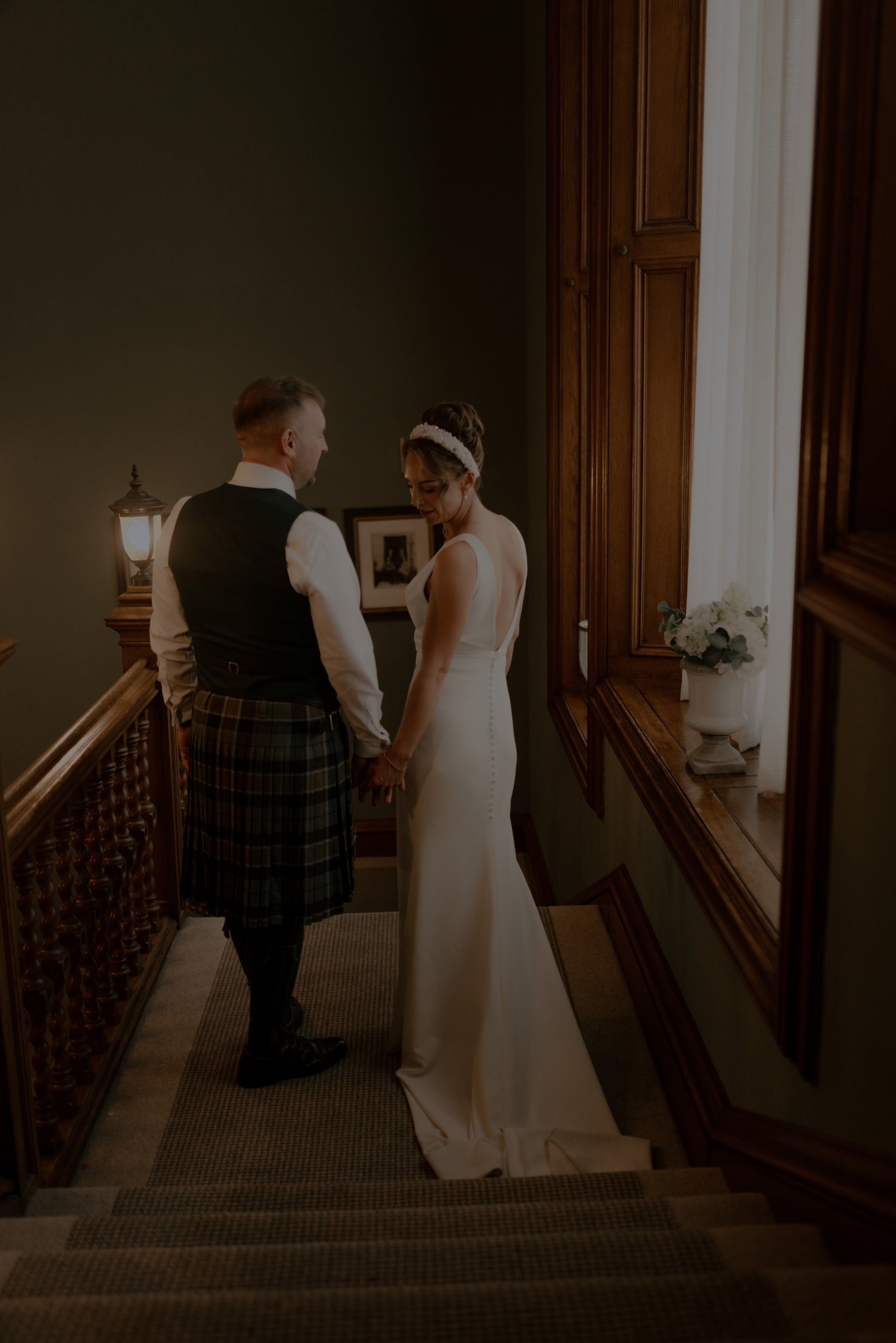 A bride and groom holding hands on staircase, facing each other, in a warmly lit, wood-paneled setting.