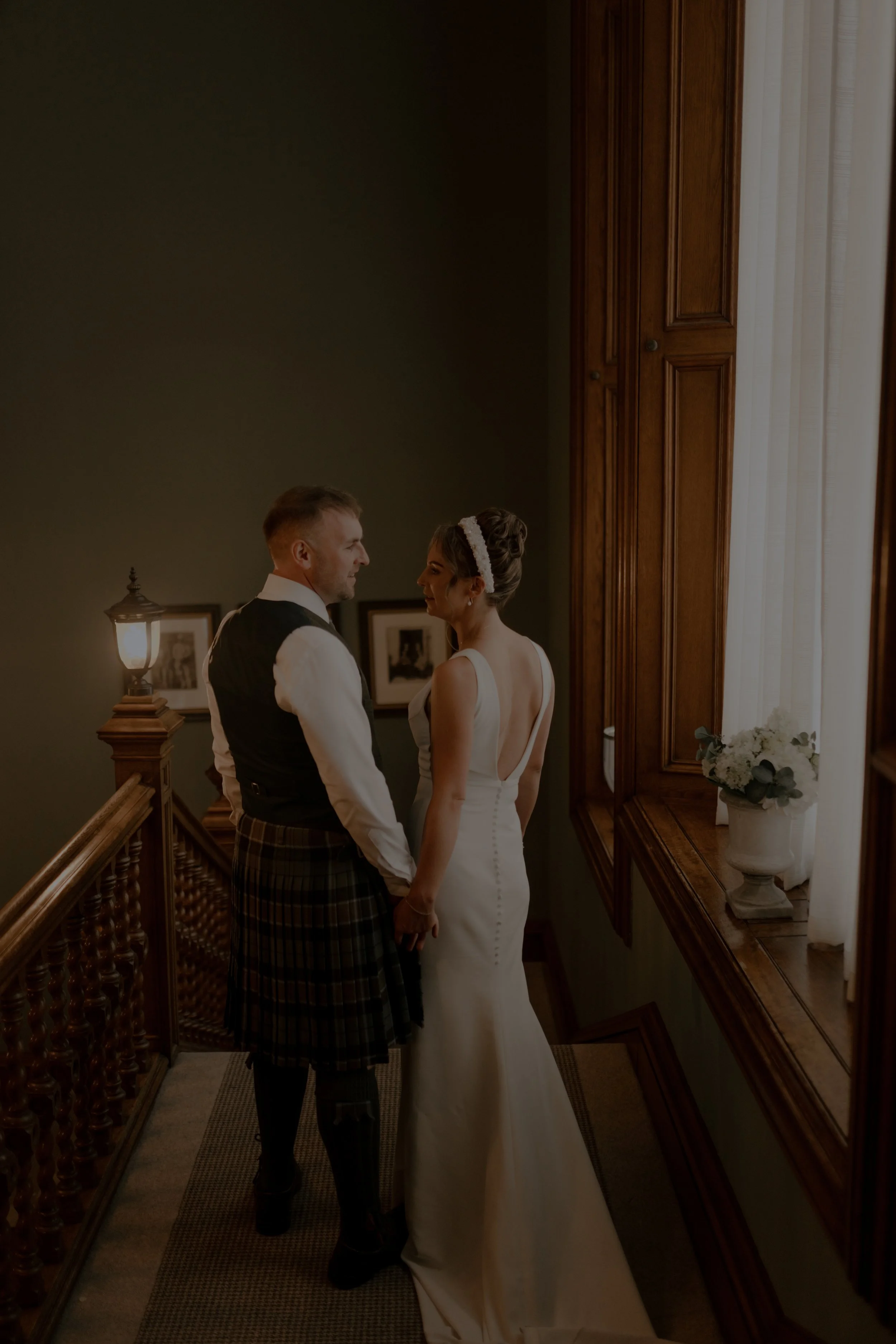 A bride and groom holding hands on a staircase at their wedding, with the bride wearing a white gown and the groom wearing traditional Scottish attire, standing close together near a window with natural light.