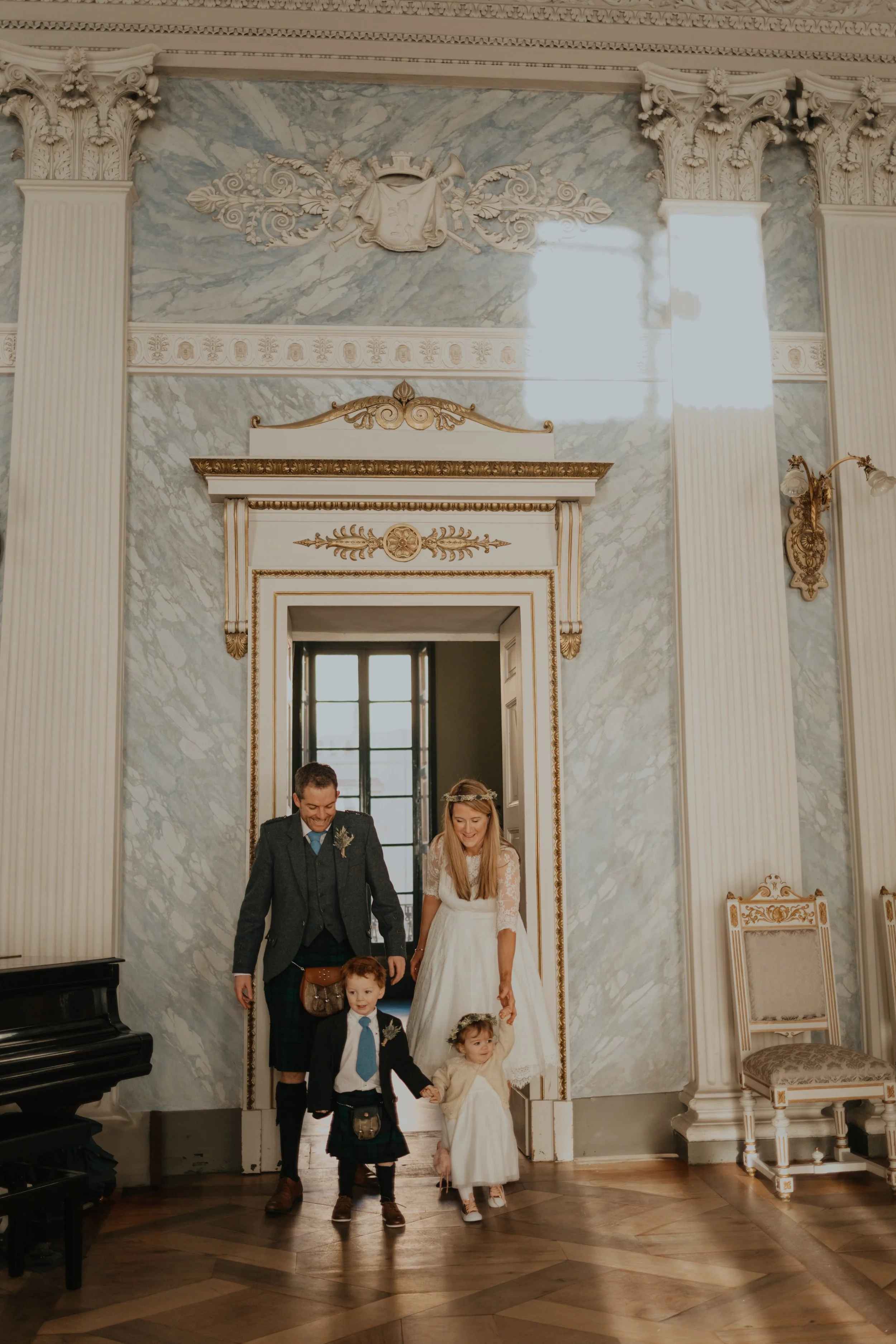 A family walking into a formal room. Two children, a boy and a girl, are dressed in suits and dresses, and a man and woman are accompanying them. The room has ornate decorations, marble walls, and wood flooring.
