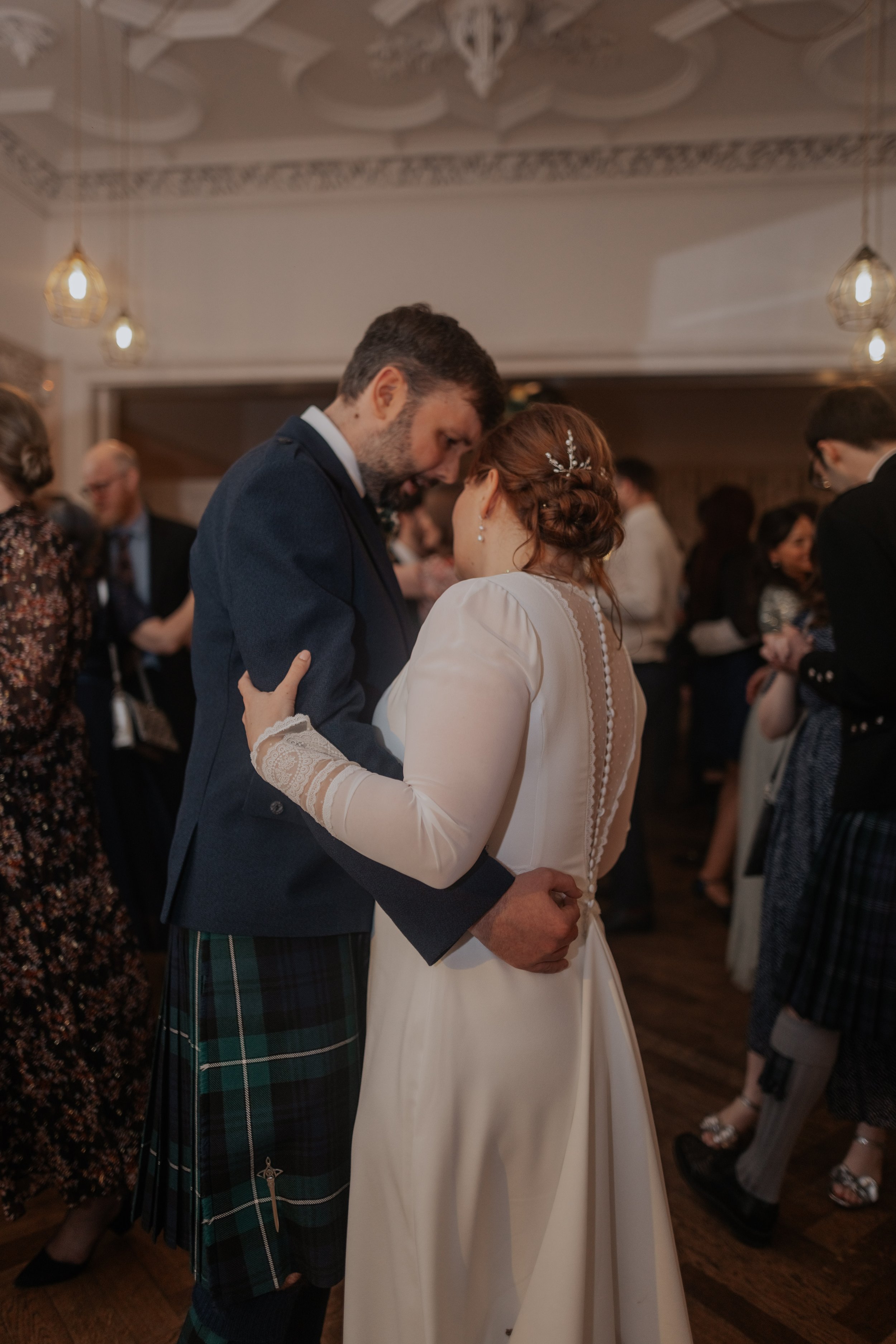 A bride and groom sharing a dance at their wedding reception, surrounded by guests in a warmly lit, elegant room.