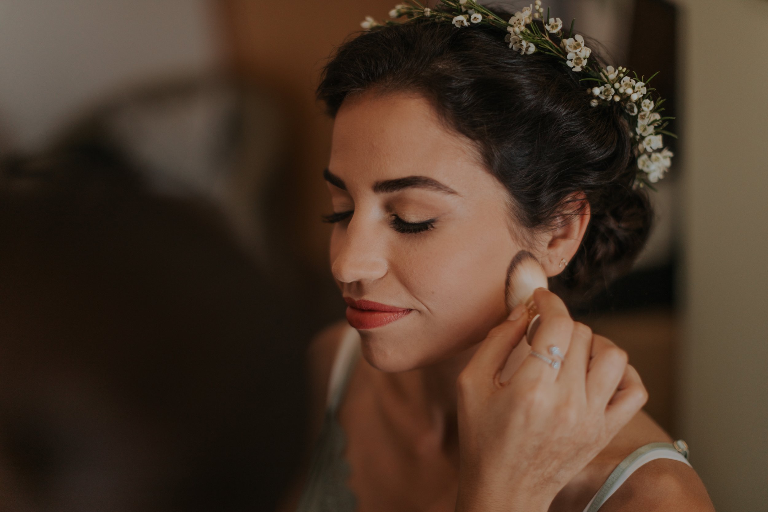 Woman with floral headpiece having makeup applied with a brush, with closed eyes and a slight smile.