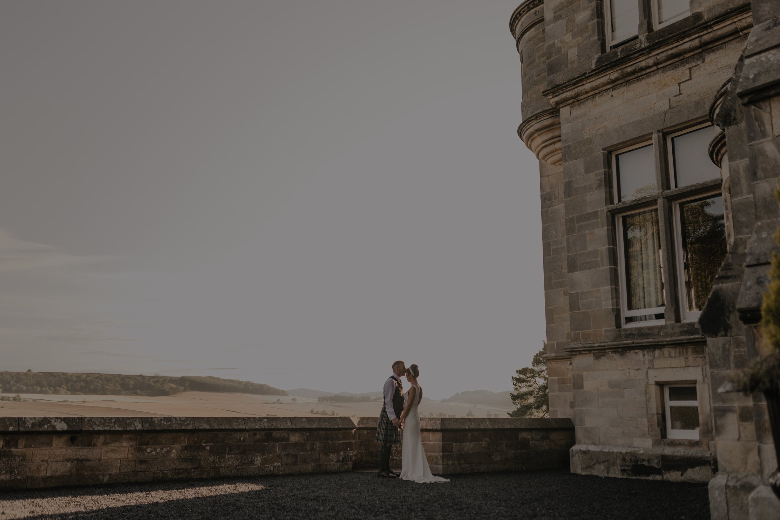 Couple in wedding attire holding hands, sharing a kiss on a balcony with a historic stone building, overlooking a rural landscape during sunset.