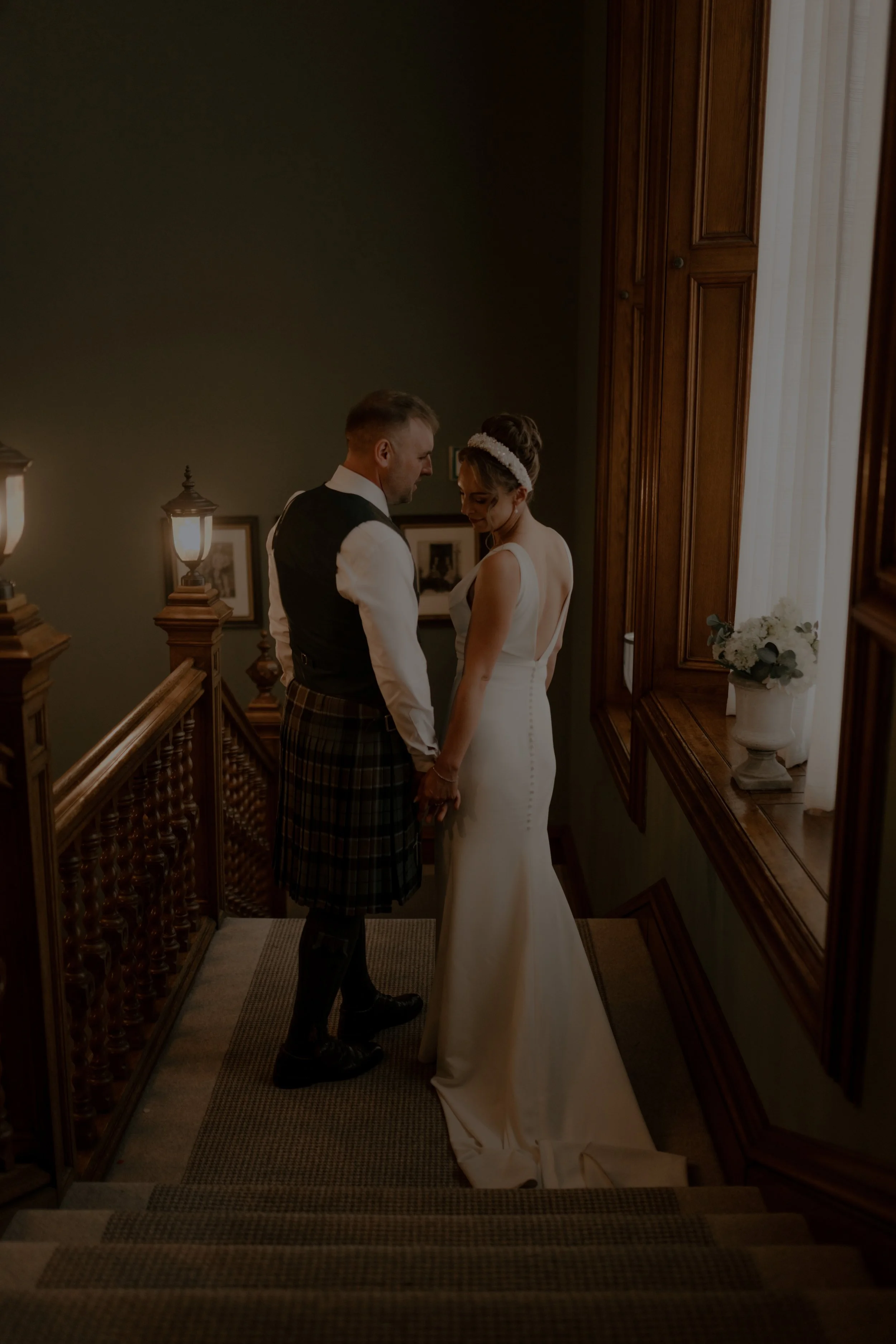 A bride and groom holding hands on a staircase, standing close together, in a dimly lit elegant home setting.