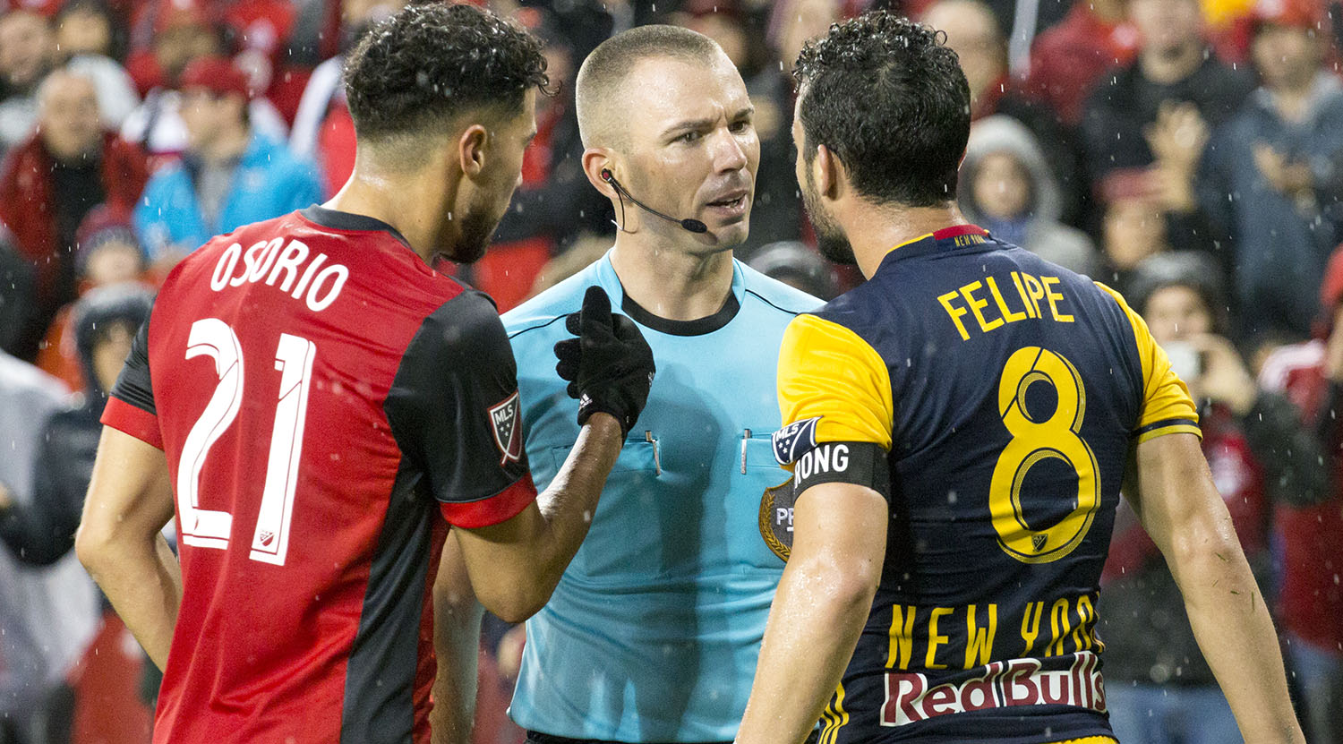 A Toronto FC and New York Red Bull player argue with the first official Chris Penso as he explains a call to both players. Image by Dennis Marciniak of denMAR Media.