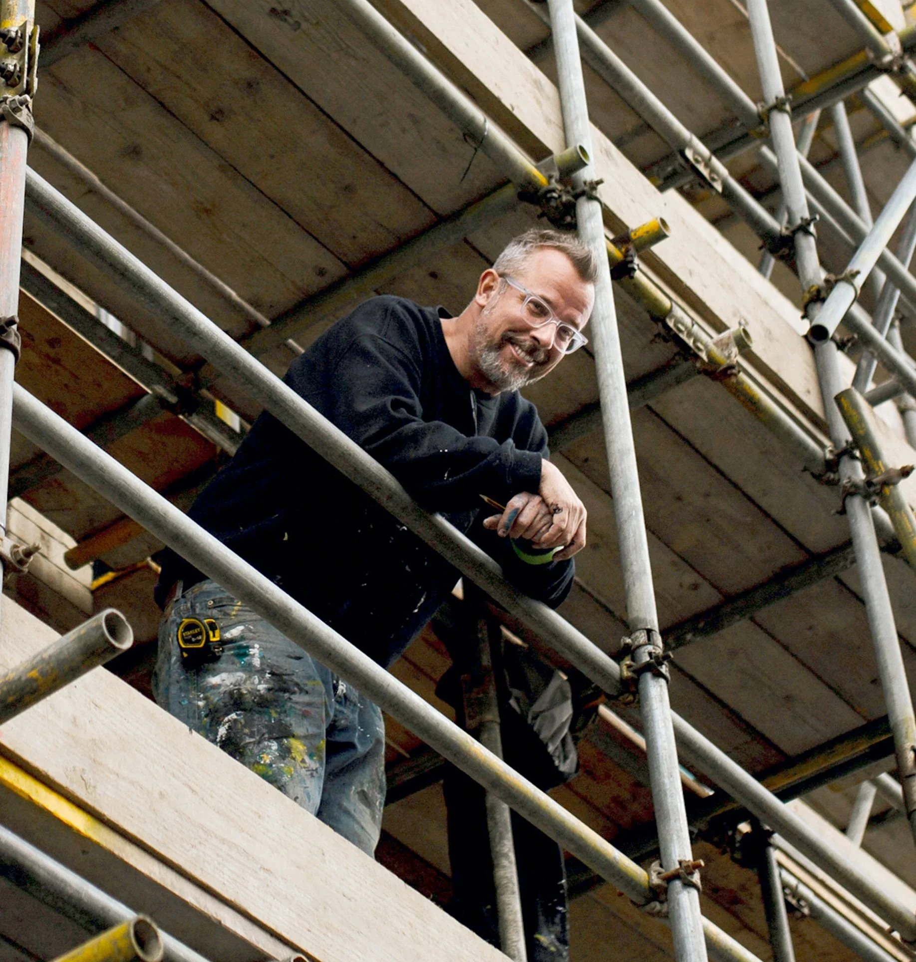 A man with glasses and a beard smiling while leaning on scaffolding at a construction site, wearing a black long-sleeve shirt and paint-splattered jeans.