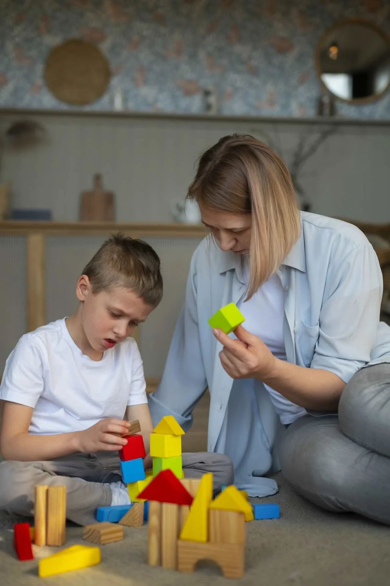 A woman and a boy playing with colorful wooden building blocks on the floor in a cozy living room.