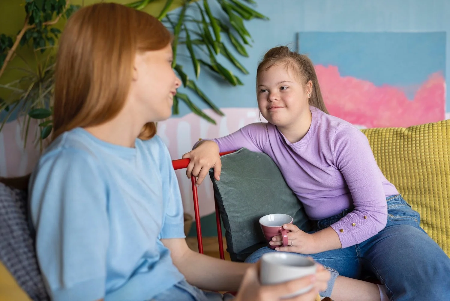 Two young girls having a conversation while sitting on a colorful couch in a cozy living room, holding mugs.