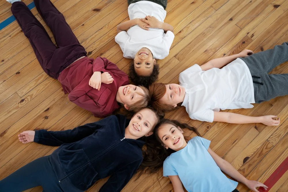 Five children lying on a wooden floor in a circle, smiling and looking up at the camera.